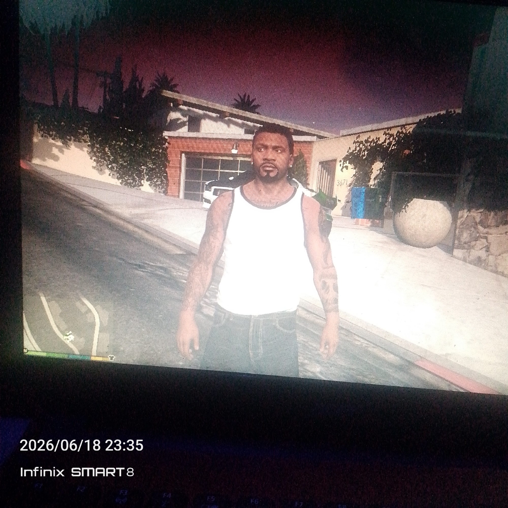 A male, appearing to be in his late 20s or early 30s, stands outdoors in front of a house. He has short, dark hair and a goatee. He is wearing a white tank top and blue jeans. The man has tattoos on his arms and chest. The house in the background has a brick facade and a pitched roof. A palm tree is visible to the left of the house, and a large, round planter sits on the driveway to the right. The sky above is a deep red and purple, suggesting either sunset or sunrise. The scene is captured from a slightly low angle, with a portion of a digital display visible at the bottom of the image, showing "2026/06/18 23:35" and "Infinix SMART 8".