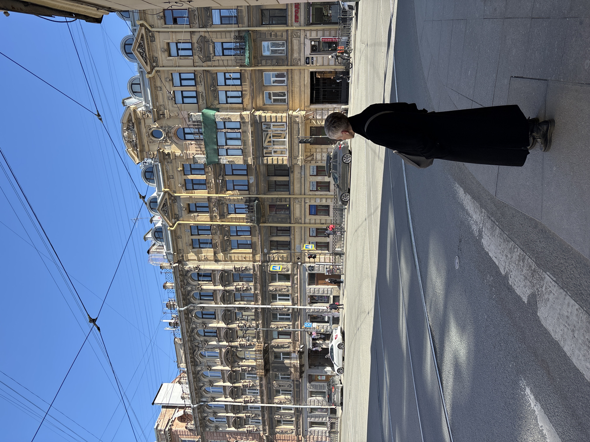 The image shows a street scene in Saint Petersburg, Russia. A tall, ornate building with multiple floors and arched windows dominates the left side of the frame. Electrical wires are visible against a clear blue sky. On the right, a woman in a long black coat stands on the sidewalk. A grey handbag hangs off her shoulder. The sidewalk and street show typical urban textures. A car is parked along the street. The composition is somewhat tilted, suggesting the photo was taken at an angle. The scene is bathed in sunlight, casting shadows on the street and sidewalk.