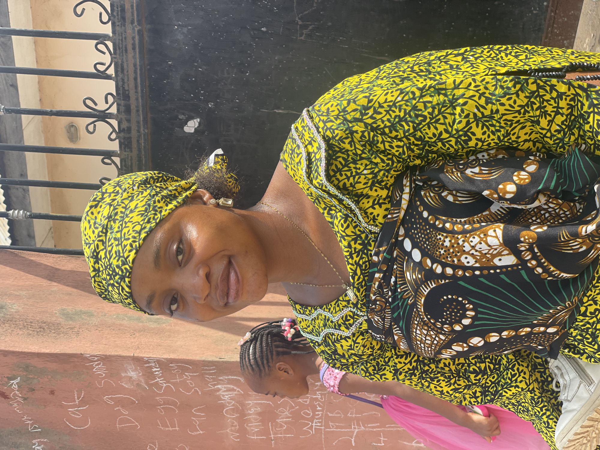 A woman in a yellow-patterned headscarf and matching top is standing next to a young girl with braided hair. The woman is smiling at the camera and carrying the young girl in front of her, wrapped in patterned cloth. A wall with writing on it is behind them. A wrought-iron gate is visible above the woman's head. The image appears to be taken outdoors, possibly in a residential area of Lokoja, Nigeria.