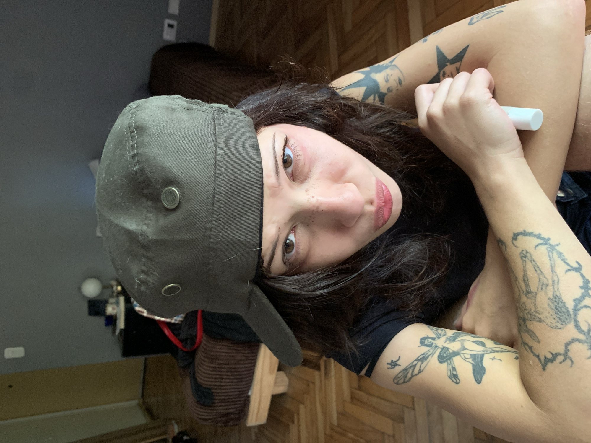 The image is a close-up, tilted portrait of a young individual, likely a selfie, rotated 90 degrees clockwise. The person has long, dark, wavy hair, light brown eyes, and prominent freckles scattered across their nose and upper cheeks. Their lips are rosy, and they engage the camera with a direct gaze. They wear a dark olive green baseball cap and a dark t-shirt. Visible on their arms are several tattoos, including a winged figure on one forearm, and star-like and other abstract designs on the other, along with a possible insect or turtle tattoo. In their right hand, they hold a small, white, cylindrical object, resembling lip balm.
The setting is indoors, featuring a partial view of a dark brown sofa or chair against a plain grey wall. A wooden parquet floor is visible, contributing to the indoor ambiance. The lighting is soft and even. No specific text is visible in the image. The scene suggests a casual, personal moment within a home environment, consistent with an urban dwelling, for example, in Buenos Aires, Argentina.