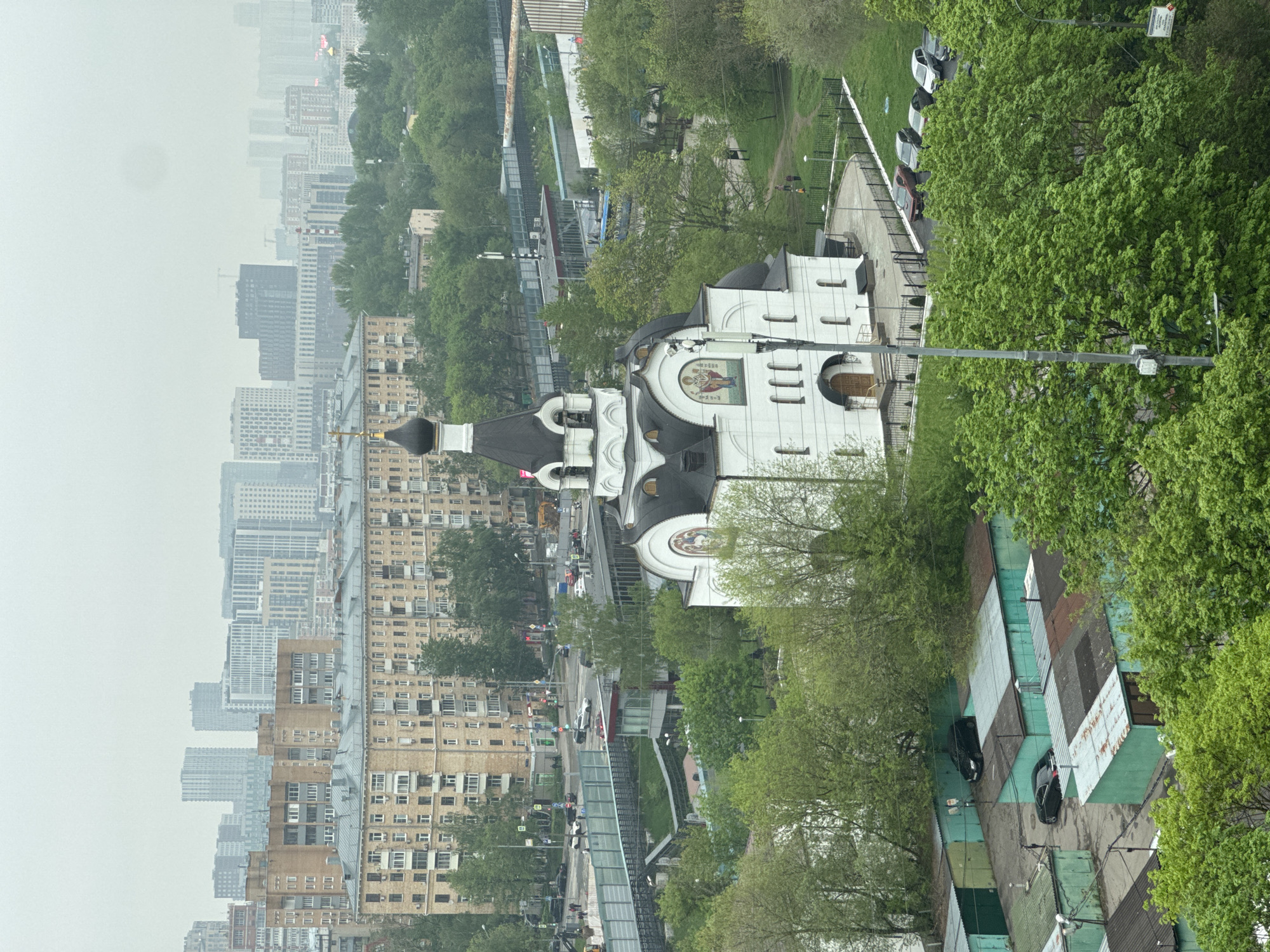The image shows an elevated view of a cityscape, likely an urban area. In the foreground, there is a white building with dark domes, possibly a church or cathedral, with intricate architectural details. This building is surrounded by green trees and foliage, suggesting a park-like setting. Beyond the trees and the church, there are rows of apartment buildings, varying in height and color, indicating different eras of construction. In the background, there is a dense skyline with numerous skyscrapers, hinting at a bustling city center. The sky is overcast, creating a somewhat muted color palette for the entire scene. Some streets and walkways with people and cars are visible within the buildings and trees. A series of garages is observed on the bottom-right. The location appears to be in the city of Unknown, Unknown.