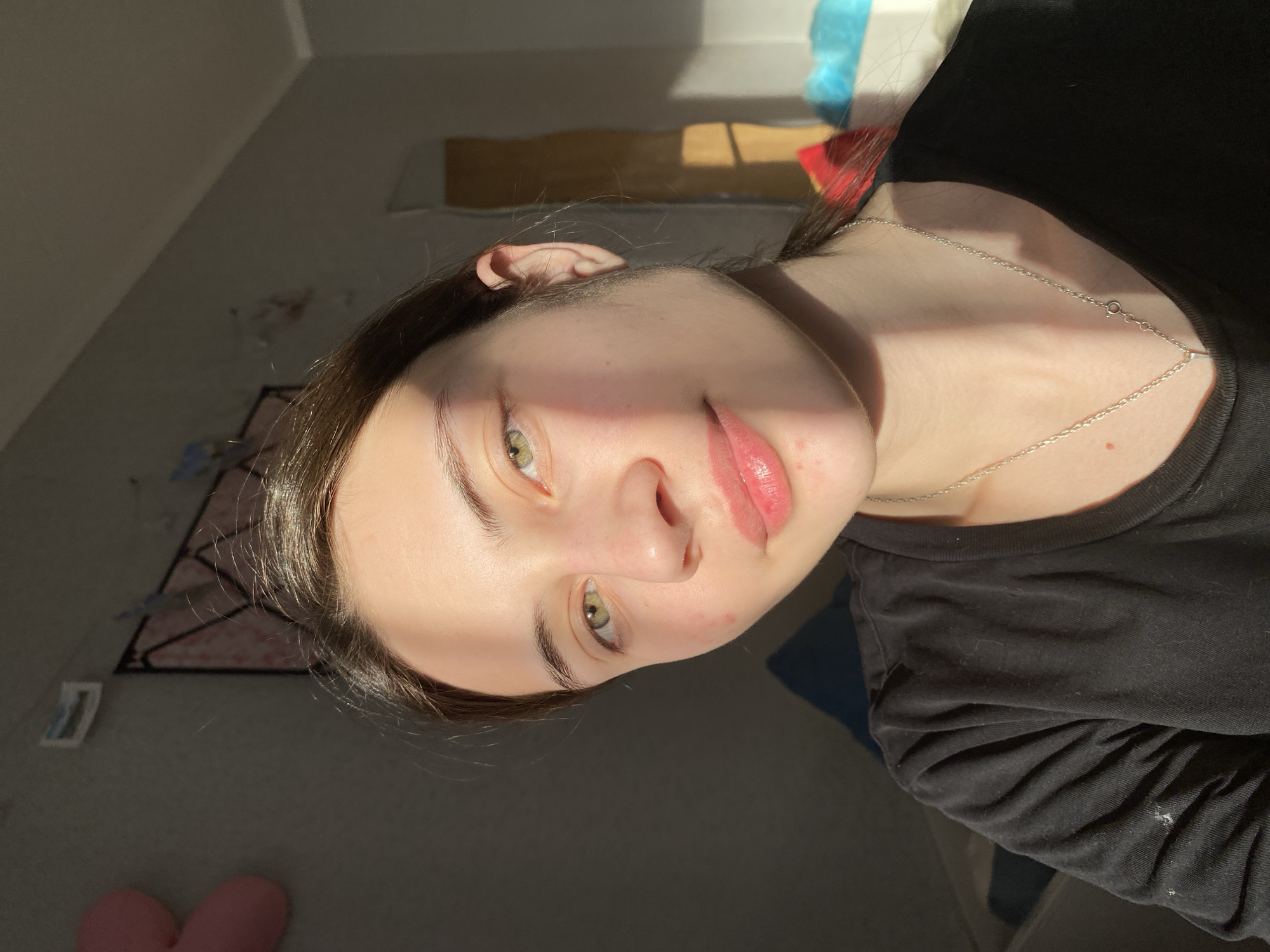 A close-up portrait of a young person with fair skin, light eyes, and dark hair. They are wearing a silver chain necklace and a black shirt. Part of their face and upper body are illuminated by strong sunlight, while the background is in shadow. The surroundings appear to be a bedroom with a bed and wall decorations. There is no verifiable information to specify the location as Votkinsk, Russia based solely on the image.