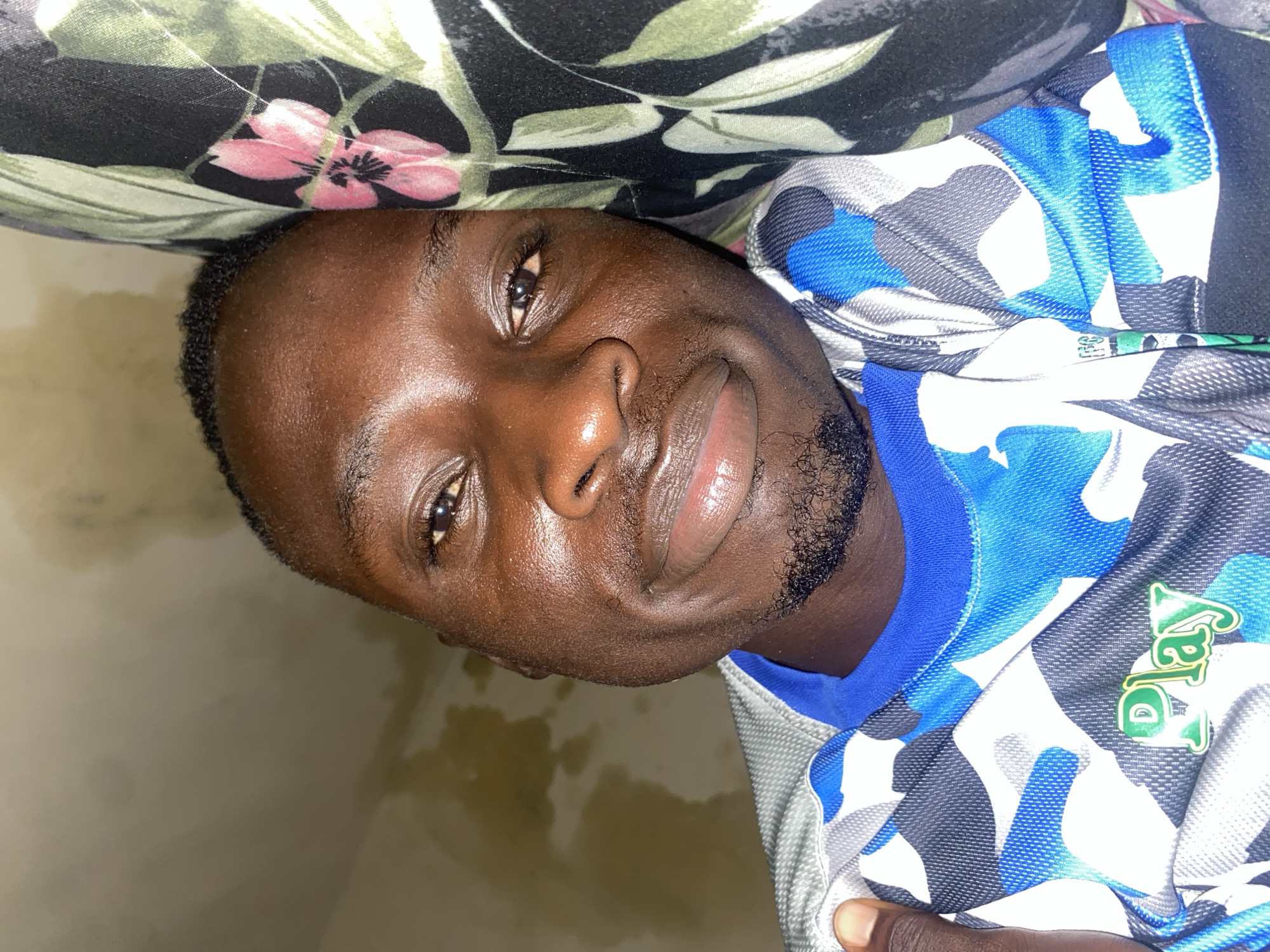 In an indoor setting, a dark-skinned male with short, dark hair and a light beard is shown in a close-up, sideways portrait. He is wearing a blue and gray camouflage-patterned shirt with green lettering that appears to spell "Play." His head is tilted, and he is looking towards the camera with a slight smile. Behind him, a pillow with a floral pattern featuring pink flowers and green leaves is visible. The lighting is somewhat dim, casting subtle shadows on his face. The overall scene suggests a casual, relaxed moment captured indoors, possibly in a bedroom or living area in Barden Yana, Nigeria.
