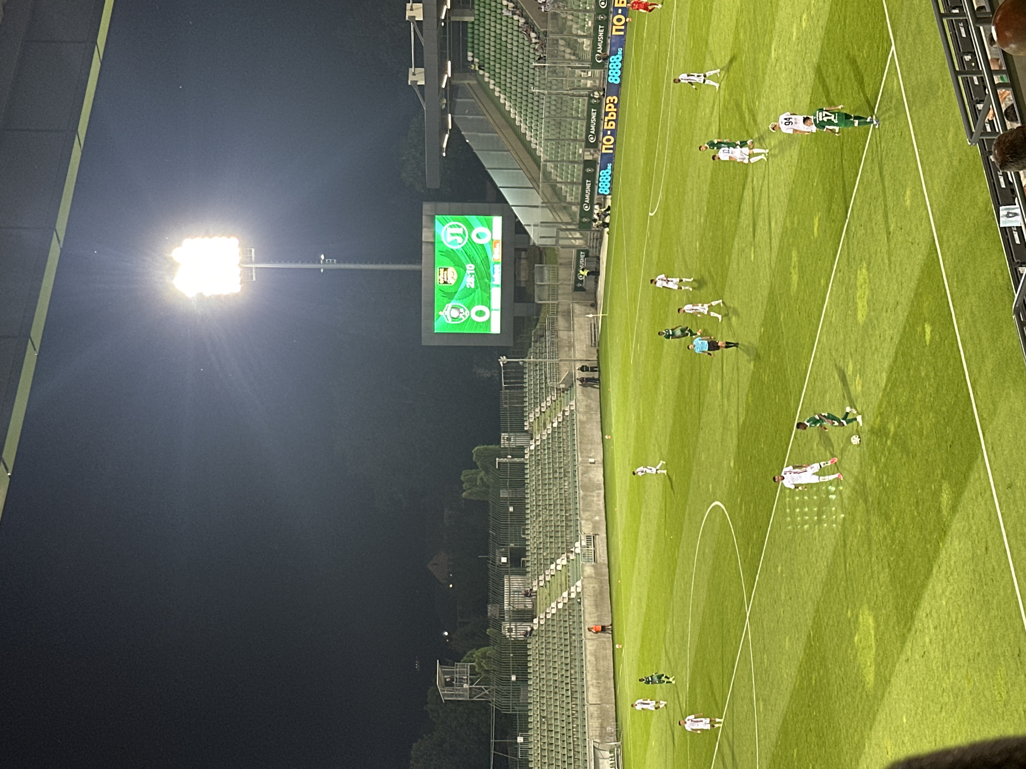 The image shows a night-time view of a football (soccer) match at the Huvepharma Arena in Razgrad, Bulgaria. The stadium is illuminated by bright floodlights. In the foreground, several football players in white and green kits are visible on the lush green pitch, engaged in the game. A large video screen is prominently displayed in the background, showing the match score and time. Spectator stands, mostly empty, are visible on the left and right sides of the stadium. The overall scene depicts a typical professional football match environment.
