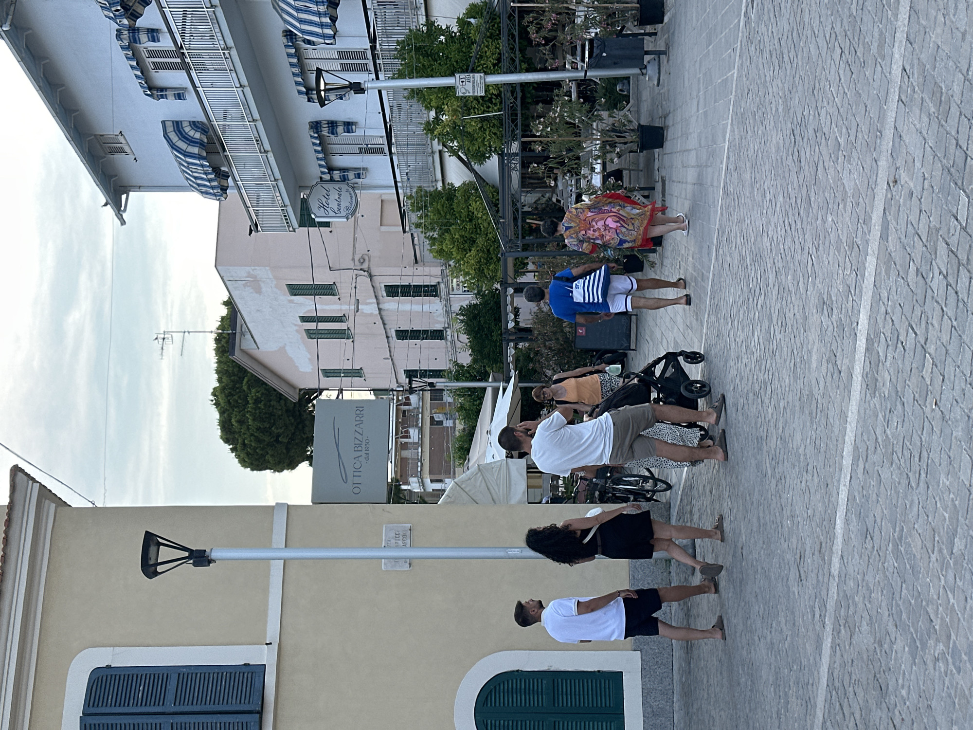 In Pineto, Italy, the image captures a street scene with several people walking on a cobblestone sidewalk. A group of four individuals, two men and two women, are in the foreground, with two of them appearing to be a couple. One man is wearing a white polo shirt and black shorts, while the woman beside him has a black dress. Further ahead, a man in a white t-shirt and khaki shorts is walking with a baby stroller. A woman in an orange tank top and patterned shorts is also present with the stroller. In the background, there are buildings with awnings, one of which is labeled "Hotel Centrale Ristorante." Another sign reads "OTTICA BIZZARRI - dal 1950". The environment suggests a commercial or residential area with shops and cafes. The sky is overcast, indicating a cloudy day.