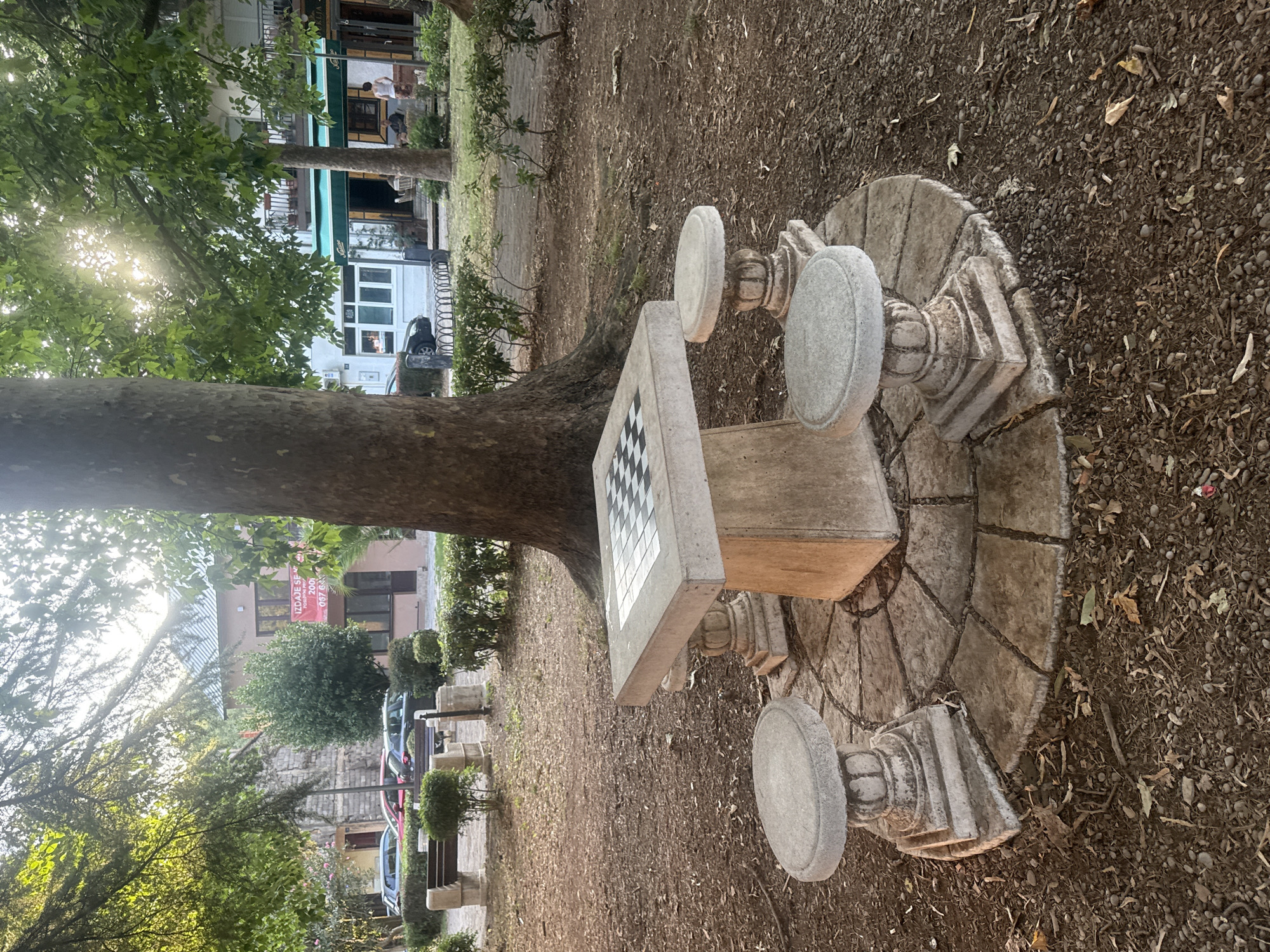 This is a full, eye-level, outdoor shot of a chess table and stools in the city of Virpazar, Montenegro. The chess table is made of concrete with a chessboard embedded in its surface. It has a pedestal base and is surrounded by three matching concrete stools, also with pedestal bases. The chess set is located on a circular paved area made of stone tiles. The ground around the paved area is covered with dirt, dry leaves, and small stones. In the background, a large tree trunk is visible to the right, and more trees and foliage are scattered throughout the scene. Behind the trees, there are buildings with windows and balconies, and some parked cars. The sun is shining through the leaves of the trees, creating a dappled light effect. The overall environment appears to be a park or a public square.