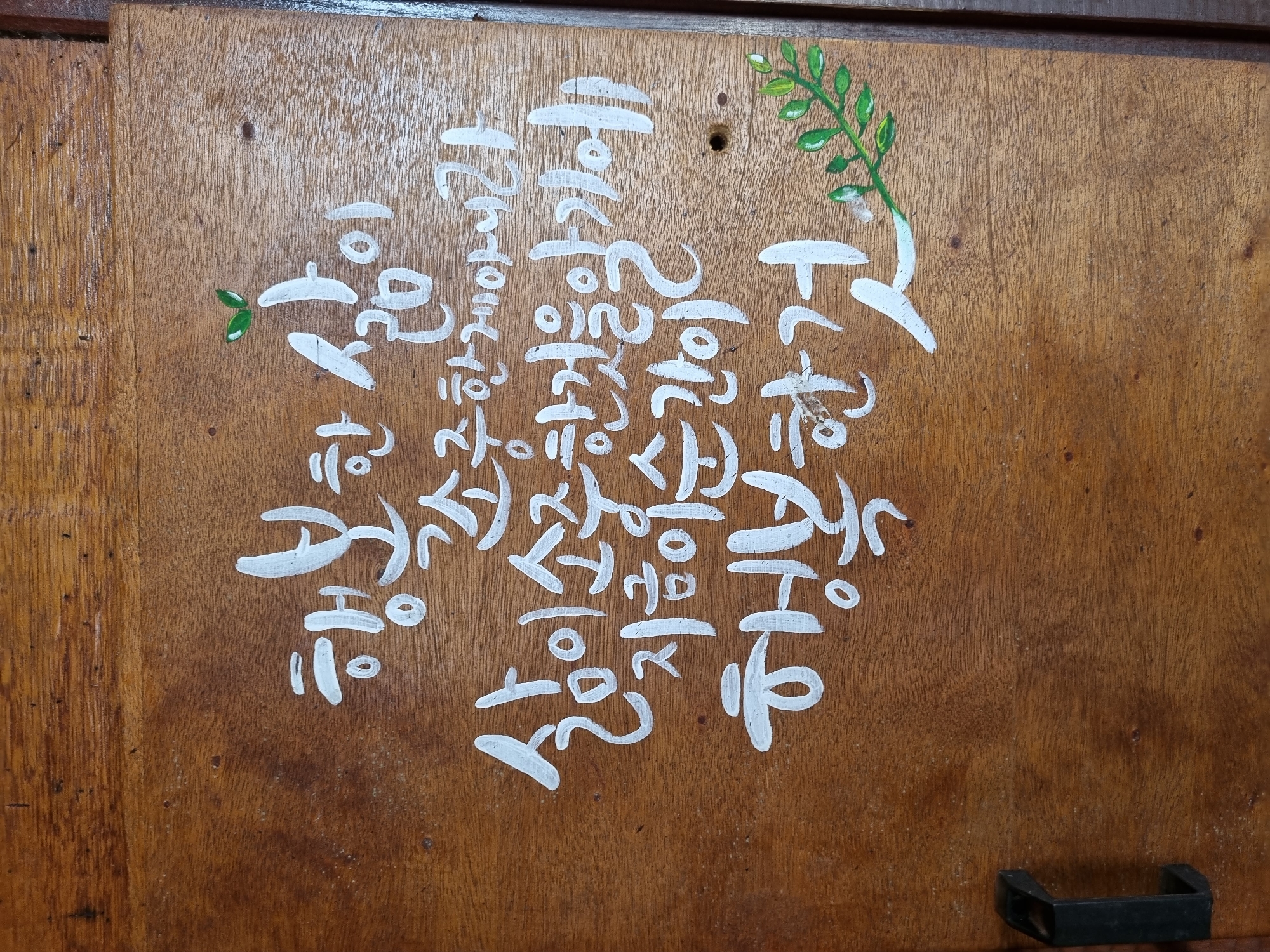The image shows white Korean calligraphy on a wooden surface. The text appears to be a quote about happiness and appreciating life. There are also small green leaves painted on the right side of the image, possibly as a decorative element. The wooden surface has visible grain and appears to be part of a piece of furniture or a wall. No people are visible in the image. The overall scene suggests a rustic or handmade aesthetic. There is no information in the image to suggest its location in Laranjal Paulista, Brazil.