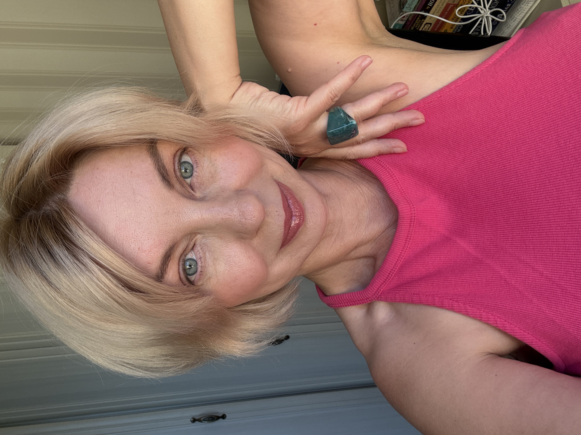 A blonde woman with blue eyes and fair skin smiles at the camera, her head tilted to the right. She is wearing a bright pink sleeveless top. Her right arm is bent and raised, with her hand resting on her head, showcasing a large, green ring on her index finger. The background consists of a pale wall with vertical lines and a closed door with a dark handle. The lighting suggests it is daytime, possibly indoors with natural light coming from the side. The woman appears to be in a personal, indoor setting. The provided image does not contain any information that would allow for the identification of a specific location in Sochi, Russia.
