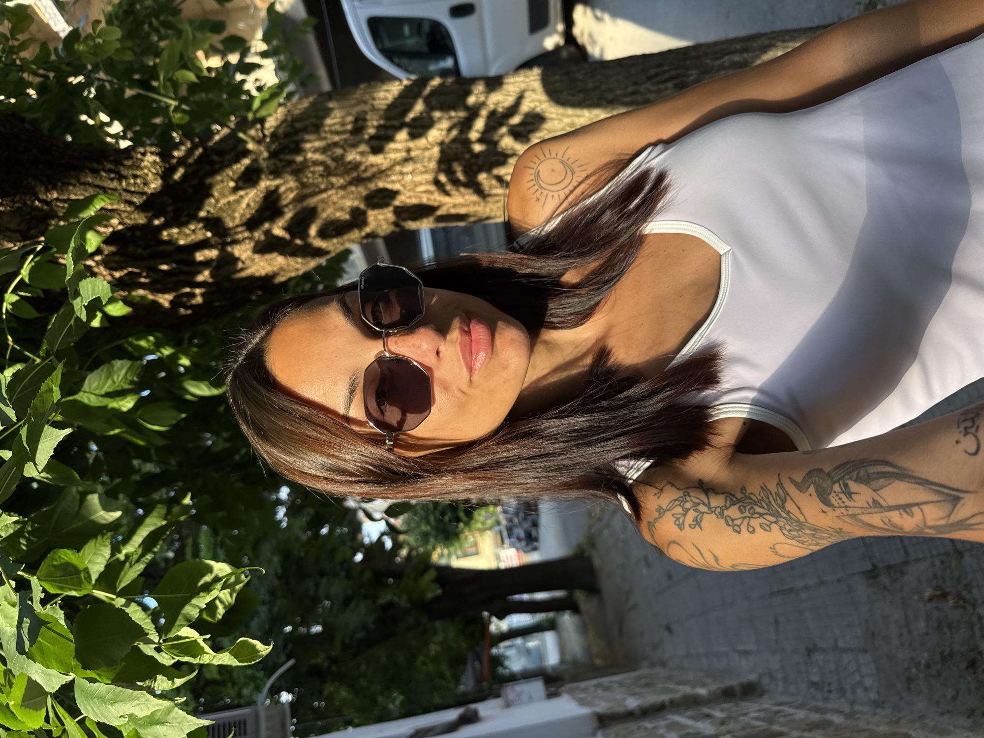 A woman with dark hair and sunglasses is posing for a selfie in Pleven, Bulgaria. She is wearing a white tank top and has several tattoos on her arms. The background shows a tree with dappled sunlight, a sidewalk, and a street with buildings and a white van. The image appears to have been taken outdoors during the day.