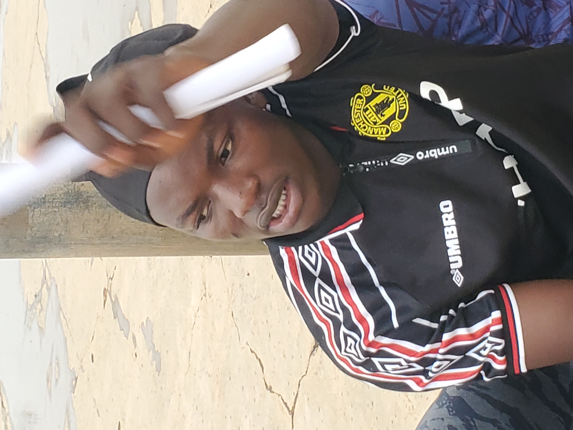 The image shows a close-up of a young person, likely male, wearing a black Manchester United jersey with red and white trim on the sleeves. The jersey has the Manchester United crest on the left chest and an "Umbro" logo on the right. The person is also wearing a black head covering. They are holding two white cylindrical objects, possibly rolled-up papers or posters, with their right hand. Their left hand is raised, and their fingers are extended towards the objects. The person's facial expression is not fully visible due to the angle and lighting, but their mouth is slightly open, revealing teeth. The background appears to be a light-colored, textured surface, possibly a wall or pavement, with some cracks and discolored patches, suggesting an outdoor setting. The lighting suggests it could be daytime. There is no clear indication of the specific time of day or weather conditions. The visible text includes "MANCHESTER UNITED" on the crest and "UMBRO" on the jersey. The setting does not provide strong location cues beyond an outdoor, possibly urban or semi-urban environment in Pankshin, Nigeria. The activity appears to be the person interacting with the cylindrical objects.