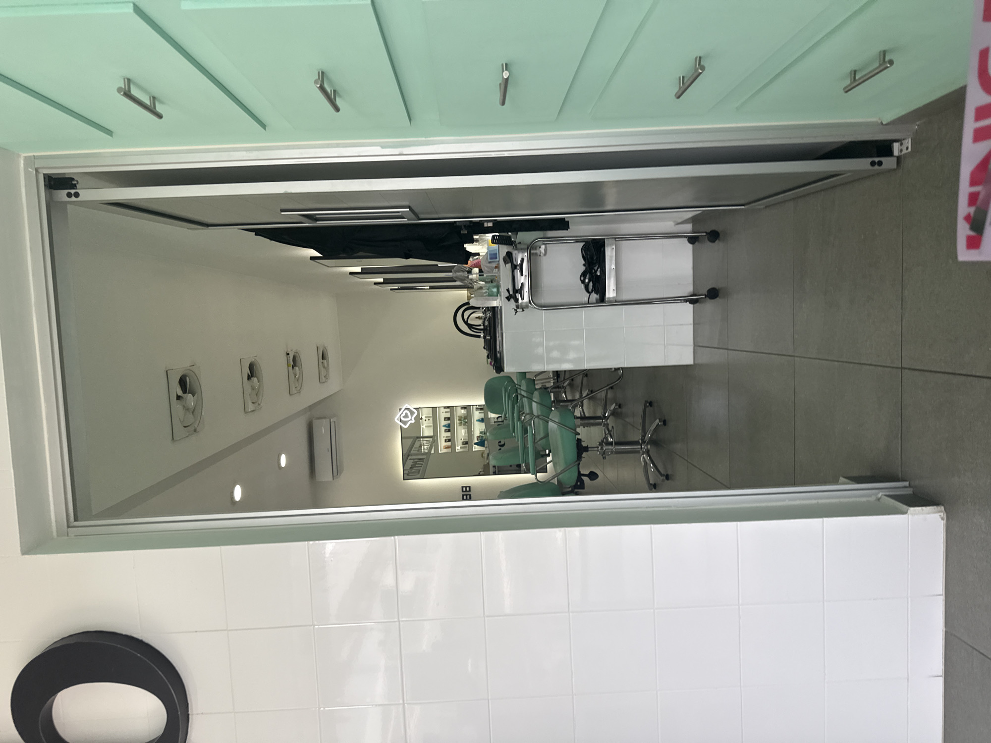 The image captures a view looking into a bright, modern salon or beauty treatment room through an open doorway. The photograph is taken from an area with light-colored flooring, possibly white tiles, which is partially visible in the bottom right corner.
To the left of the doorway, a light green wall with cabinet doors and silver handles extends upwards, suggesting overhead storage or decorative panelling in the entrance area. The doorway itself features a white or silver frame, hinting at a sliding or folding glass door mechanism.
Inside the room, the walls are predominantly white, contributing to a clean and minimalist aesthetic. Along the left wall within the salon, several square-shaped ventilation fans are mounted in a row, accompanied by a white wall-mounted air conditioning unit and a few recessed circular lights.
The main focal point inside is a large, rectangular mirror on the back wall, which reflects the room's interior. The mirror features an illuminated outline of a heart at its top center, adding a decorative touch. Reflected in the mirror are multiple light green salon chairs with chrome bases and wheels, arranged in a line, indicating several styling or treatment stations. In front of these chairs, a white workstation or counter is visible, topped with various bottles, salon products, and tools, along with electrical cords. A smaller, white rolling cart is also present near the workstation. The floor inside the salon is tiled with grey squares, which extends from the doorway.
The room appears tidy and well-maintained, suggesting a professional environment. It is unoccupied, with no people or legible text visible in the frame. The overall impression is one of a functional, clean, and inviting professional space, possibly a hair salon or beauty clinic, located in San Bernardino Tlaxcalancingo, Mexico.