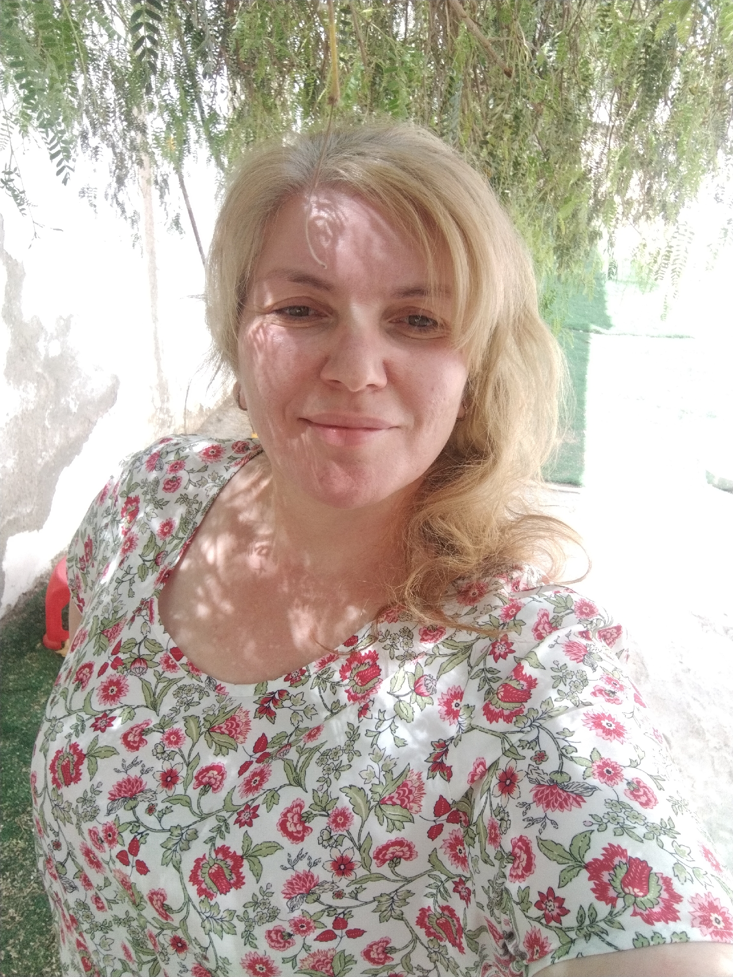 A fair-skinned, middle-aged woman with blond hair is taking a selfie. She is wearing a white floral-patterned blouse with red and green details. The background appears to be outdoors, with vegetation hanging overhead, and a white wall to her left. Based on the user's location information, the image was likely taken in Al Mas'oudi, United Arab Emirates, although there are no specific features in the image that confirms this location.