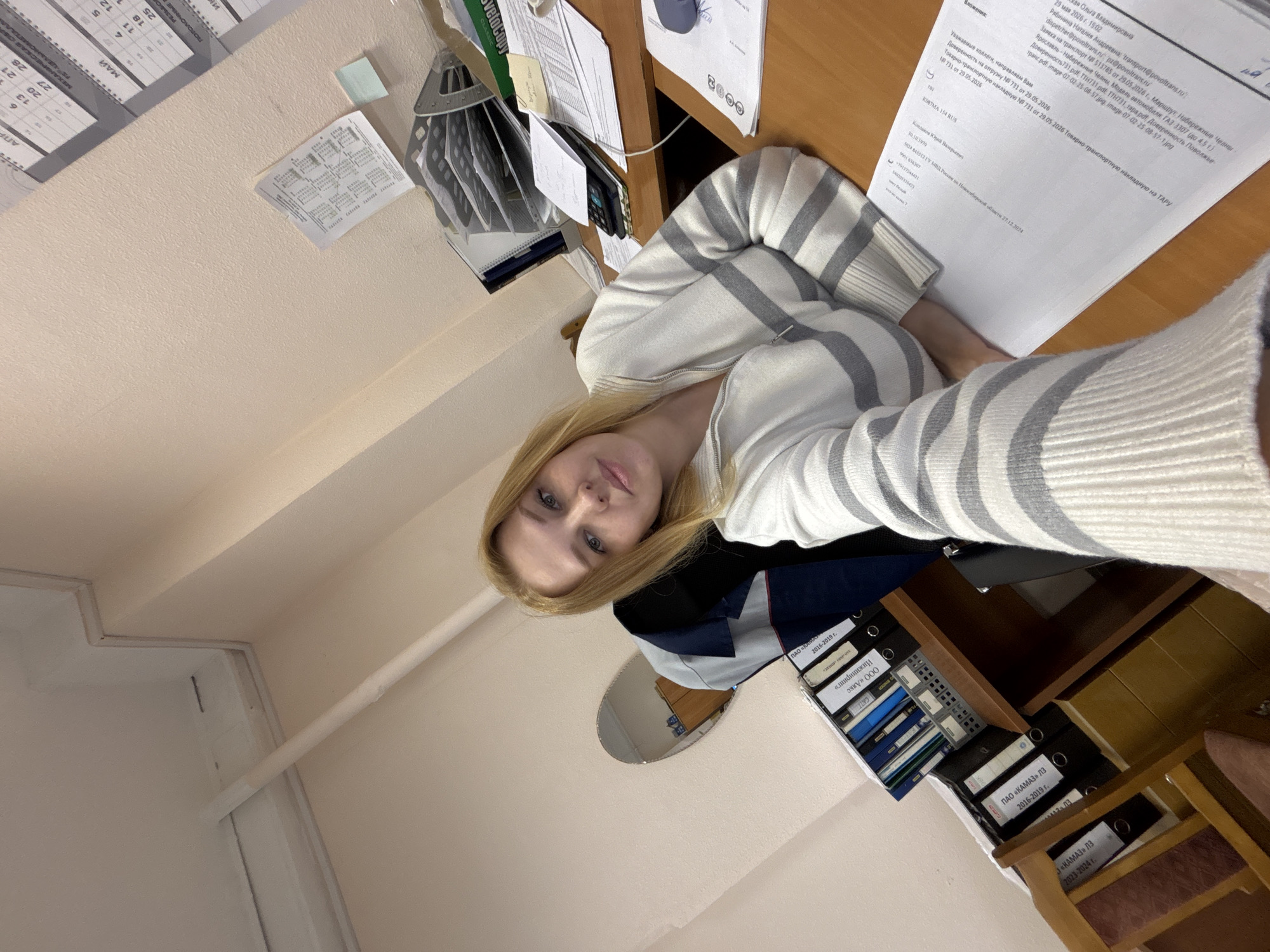 Here is a description of the image:
A woman with blonde hair is taking a selfie in what appears to be an office setting. She is wearing a striped sweater. In the background, there's a desk cluttered with papers, a calendar on the wall, and a shelf containing binders. Some of the binders have labels in Russian. The papers on the desk include what appears to be official documentation with Russian text. The lighting seems to be from overhead fluorescent lights, common in office environments. The orientation of the photo is angled, giving a slightly distorted view of the scene. Based on the text present in the image (which mentions Поволжье) and the Russian on the documents, it's likely this photo was taken in Russia. The documents also mention "Заявка на транспорт" which could translate to "transport application" or "transport request", and also references "Набережные Челны" (Naberezhnye Chelny), a city in Russia, possibly indicating the origin or destination of transport. Based on the context, the location could be an office related to transportation or logistics within Russia, but the specific city of Заволжье cannot be confirmed based on the image.