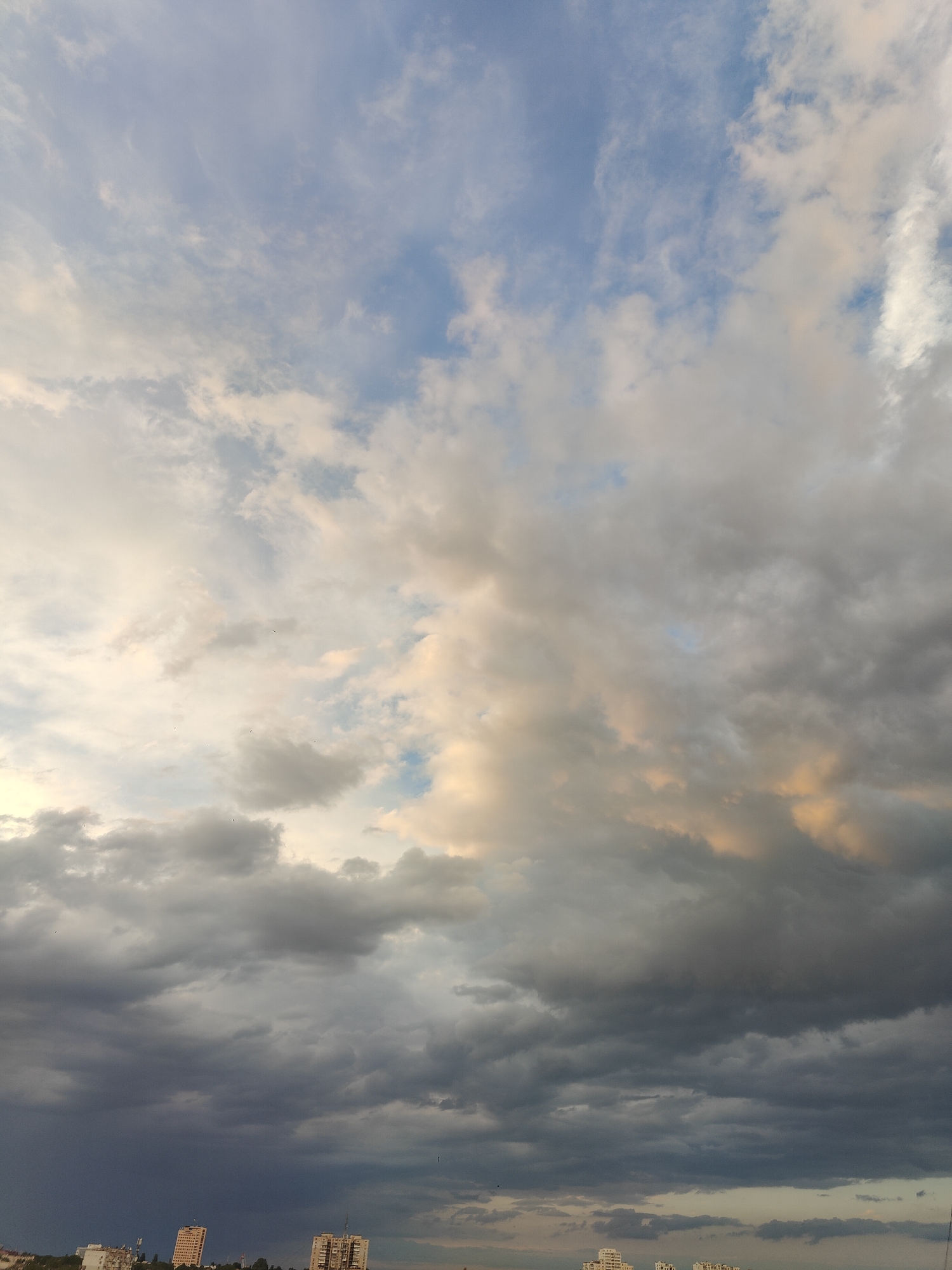 The image shows a cloudy sky above a city skyline. The clouds are varied, with some appearing dark and heavy, suggesting rain, while others are lighter and wispy. There are hints of sunlight breaking through the clouds, creating patches of yellow and orange light. In the lower part of the image, several buildings are visible, including apartment blocks and what appear to be telecommunication towers. The architecture suggests a location in Eastern Europe. Based on the provided context, the scene is likely the city of Chornomorsk, Ukraine. There are no people or specific actions depicted in the image.