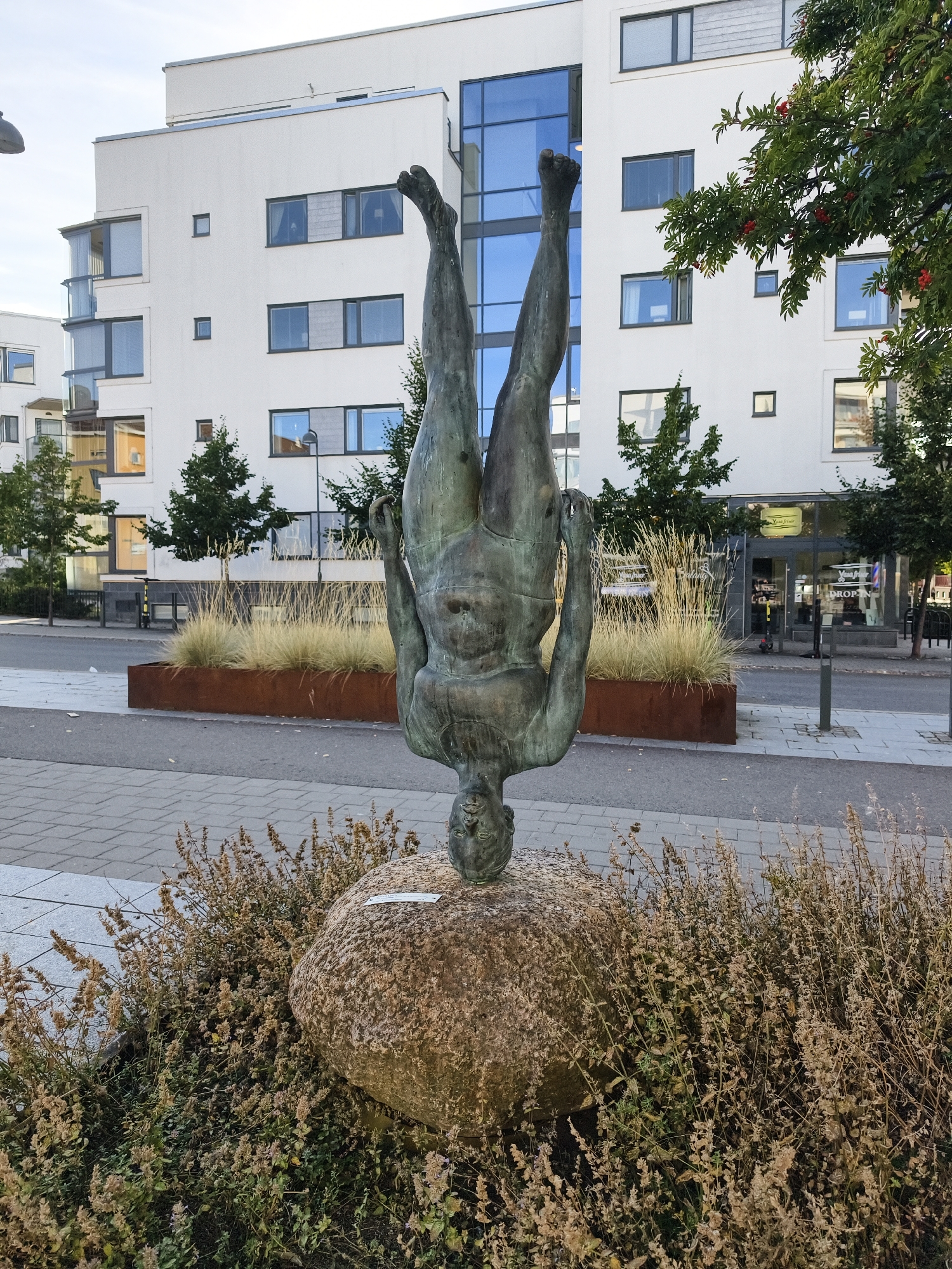 This image captures a bronze sculpture of a person in a handstand pose, balanced on their head atop a large rock. The sculpture is situated outdoors, in front of a modern, white apartment building with numerous windows and balconies. The foreground features dry, grassy plants surrounding the rock and the base of the sculpture. Behind the sculpture, a planter box with ornamental grass is visible, and beyond that, a sidewalk and a street. The building and the surrounding urban environment suggest this is a public space in a city. Based on the context provided, this scene is located at Trädgårdstorget in Linköping, Sweden, a square known for its urban art installations and modern architecture.