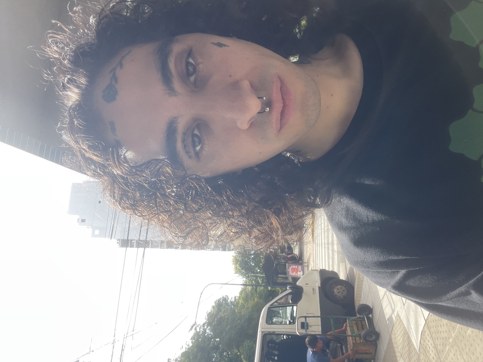A medium, angled portrait of a young man with long, curly hair, dark eyes, and multiple facial tattoos. He has a septum piercing and appears to be outdoors in an urban setting. In the background, out of focus, is a multi-story glass building, a street with parked cars, trees, a white van, and a person handling a dolly with boxes. The location is Buenos Aires, Argentina, evidenced by the architecture and style of the buildings.