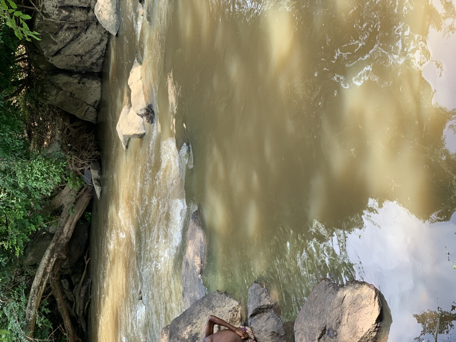 The image shows a body of water with muddy brown water, likely a river or stream. The water is moving swiftly and is slightly choppy. There are several large rocks in and around the water. One large rock in the foreground has a young person sitting on it, facing the water. The person is wearing a dark shirt and a dark skirt. The scene is in a wooded area with thick vegetation on the far bank. The scene is located at the coordinates 7.119 latitude, 5.362 longitude, in the city of Egbeda, Nigeria.
