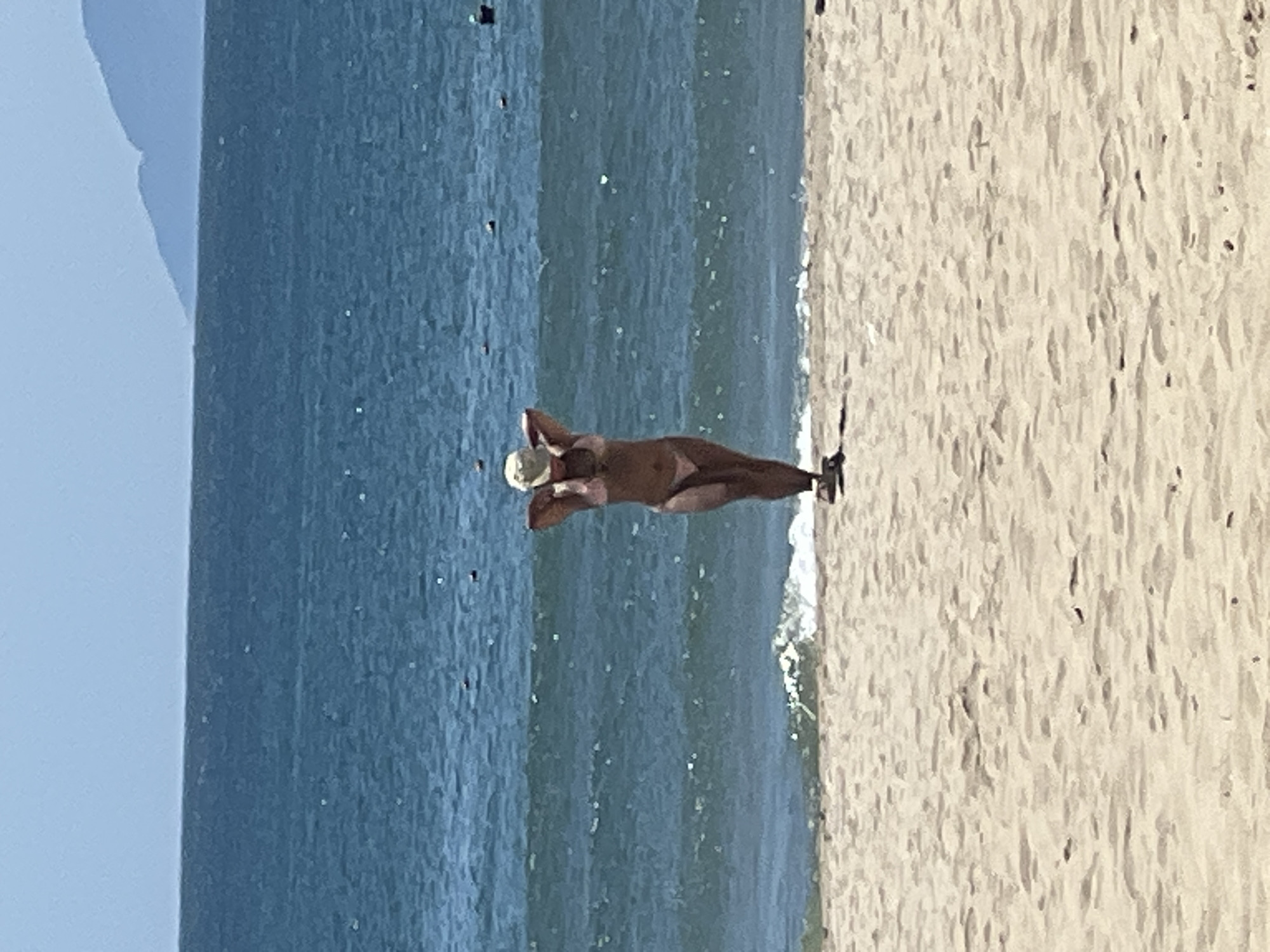 A person wearing a bikini and a hat stands on a sandy beach, facing the ocean. The water is a deep blue with some whitecaps. In the distance, hazy mountains are visible on the horizon. The scene appears to be a sunny day at a beach in Đà Nẵng, Vietnam.
