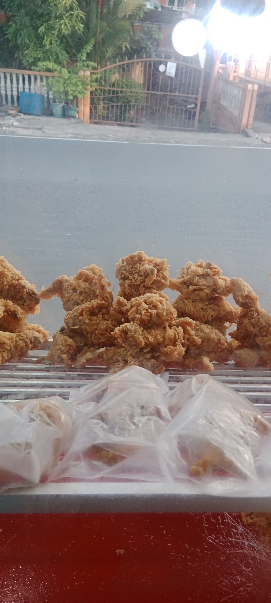 The image shows a display of fried chicken in a glass-fronted refrigerated case, likely at a food establishment in Tanjung Pinang, Indonesia. The fried chicken pieces, which appear to be whole pieces and possibly wings, are golden brown and have a crispy coating. Some of the chicken is directly on a metal rack, while other pieces are partially obscured by clear plastic packaging. The background visible through the glass shows a street scene with a metal fence, greenery, and a building with a light fixture. The overall impression is that of a street-side food vendor or a small eatery.