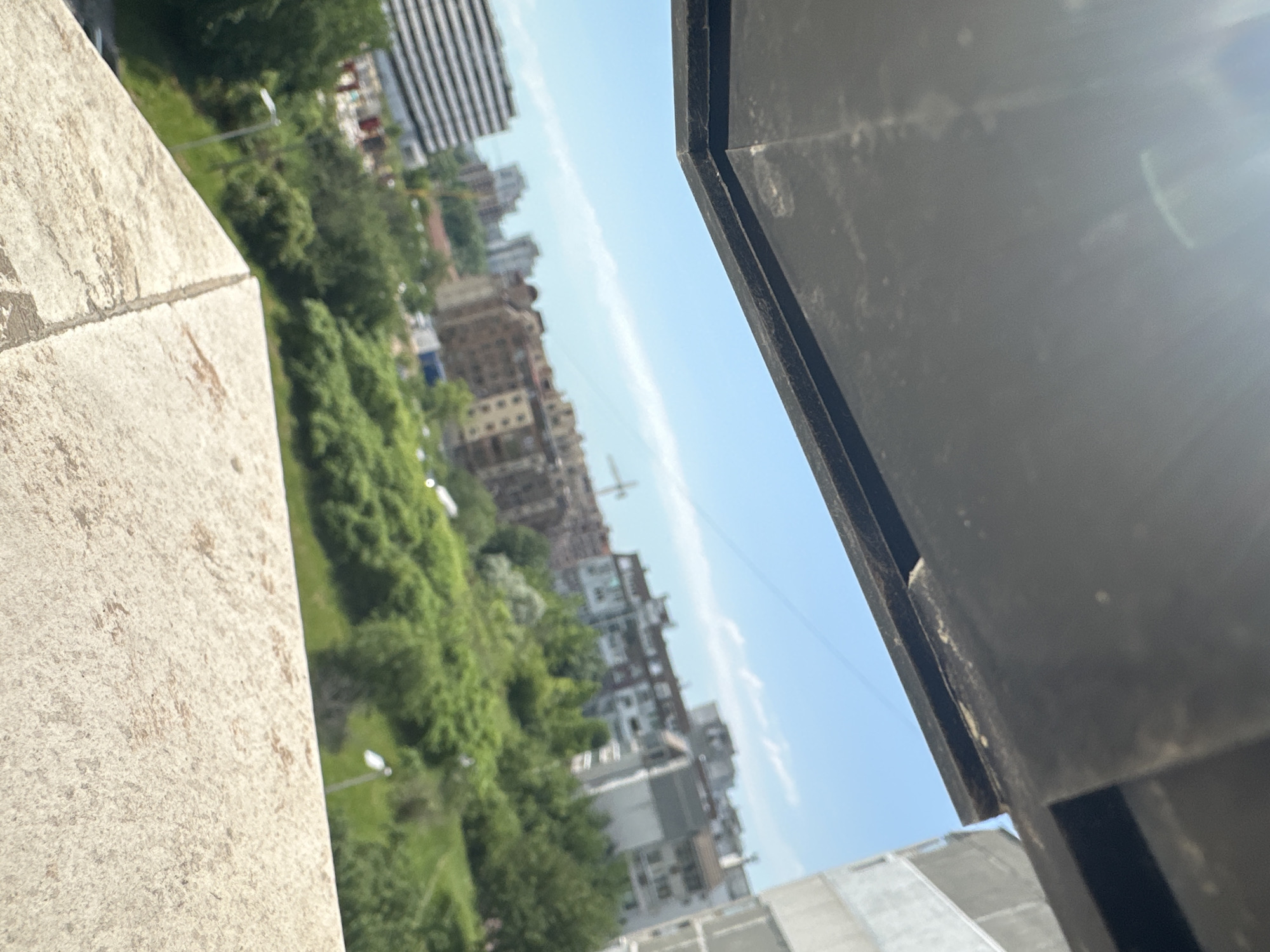 The image shows a cityscape viewed from what appears to be a high vantage point, possibly a balcony or window. In the foreground, parts of the balcony structure are visible: a section of a light-colored, textured wall on the left and a portion of a dark-colored, possibly metal or painted structure on the right. The midground consists of a dense green area, seemingly a park or forested area, interspersed with buildings of varying heights and architectural styles. Some of the buildings are modern, high-rise structures, while others appear older and of mid-rise construction. In the distance, the sky is visible, with a light blue hue and some wispy clouds. A construction crane is also visible. While specific details for confirming it's Varna, Bulgaria, are missing, the cityscape composition and general architectural styles are consistent with urban areas in Bulgaria.