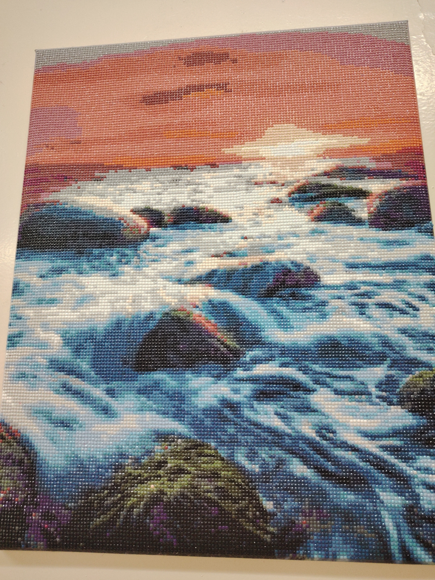 The image depicts a square-shaped craft piece. The artwork features a scene of water flowing around rocks under a sunset sky. The sky is rendered in shades of orange, pink, and grey. The water is a mixture of blues and whites. The rocks are various shades of green and black. The entire scene is composed of small, square, mosaic-like pieces. The image appears to be a completed diamond painting. There is no indication of the location in the city of Sarapul, Russia.