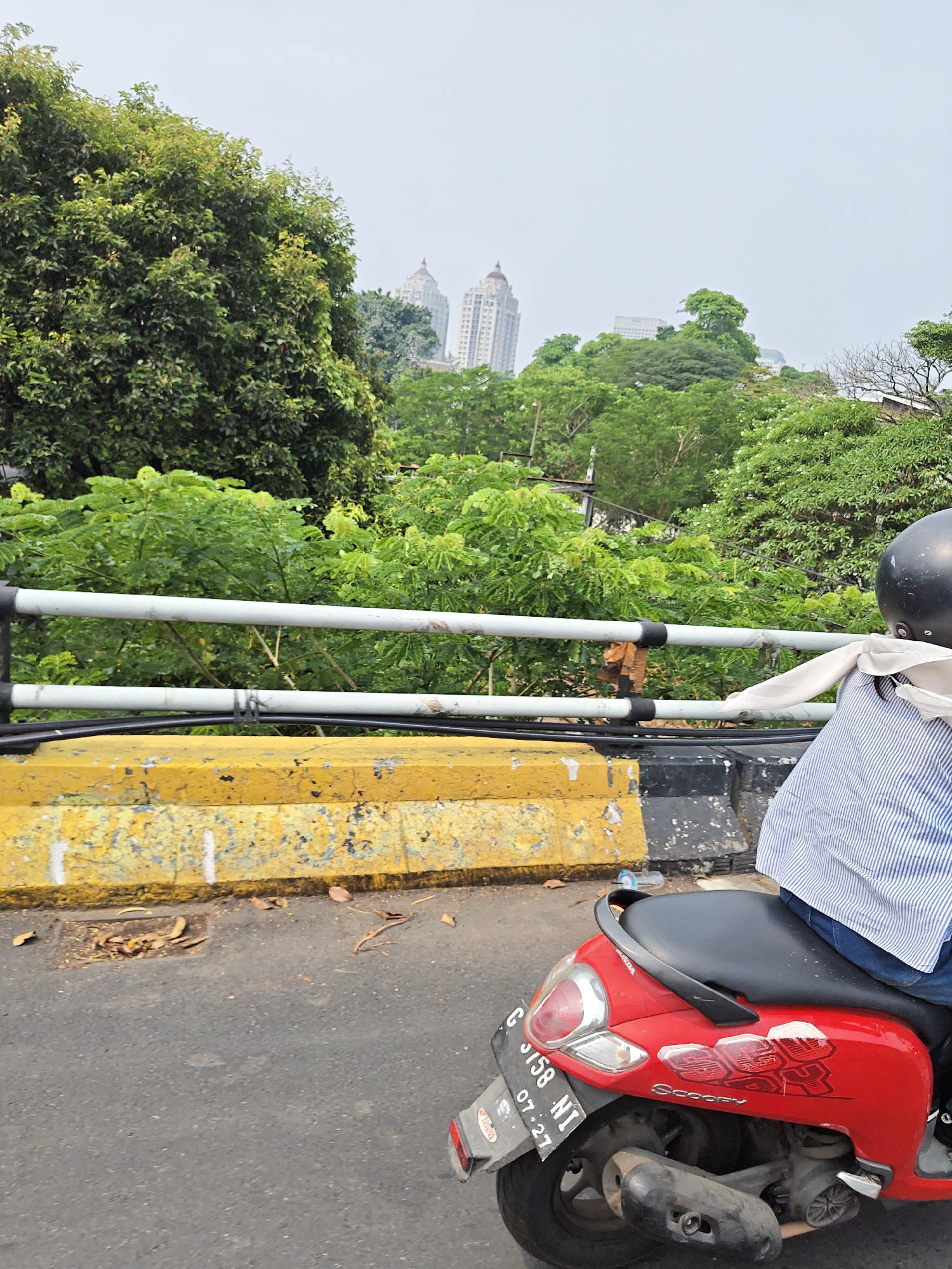A red Honda Scoopy motorcycle is parked on a street in South Jakarta, Indonesia. The motorcycle has a black seat, a black helmet on the backrest, and a blue and white striped shirt draped over the seat. A license plate reading "B 3158 NI 07-27" is attached to the rear fender. The street is paved with asphalt and has a yellow concrete barrier with black markings. Behind the barrier is a lush green landscape with trees and buildings in the distance. The sky is overcast.