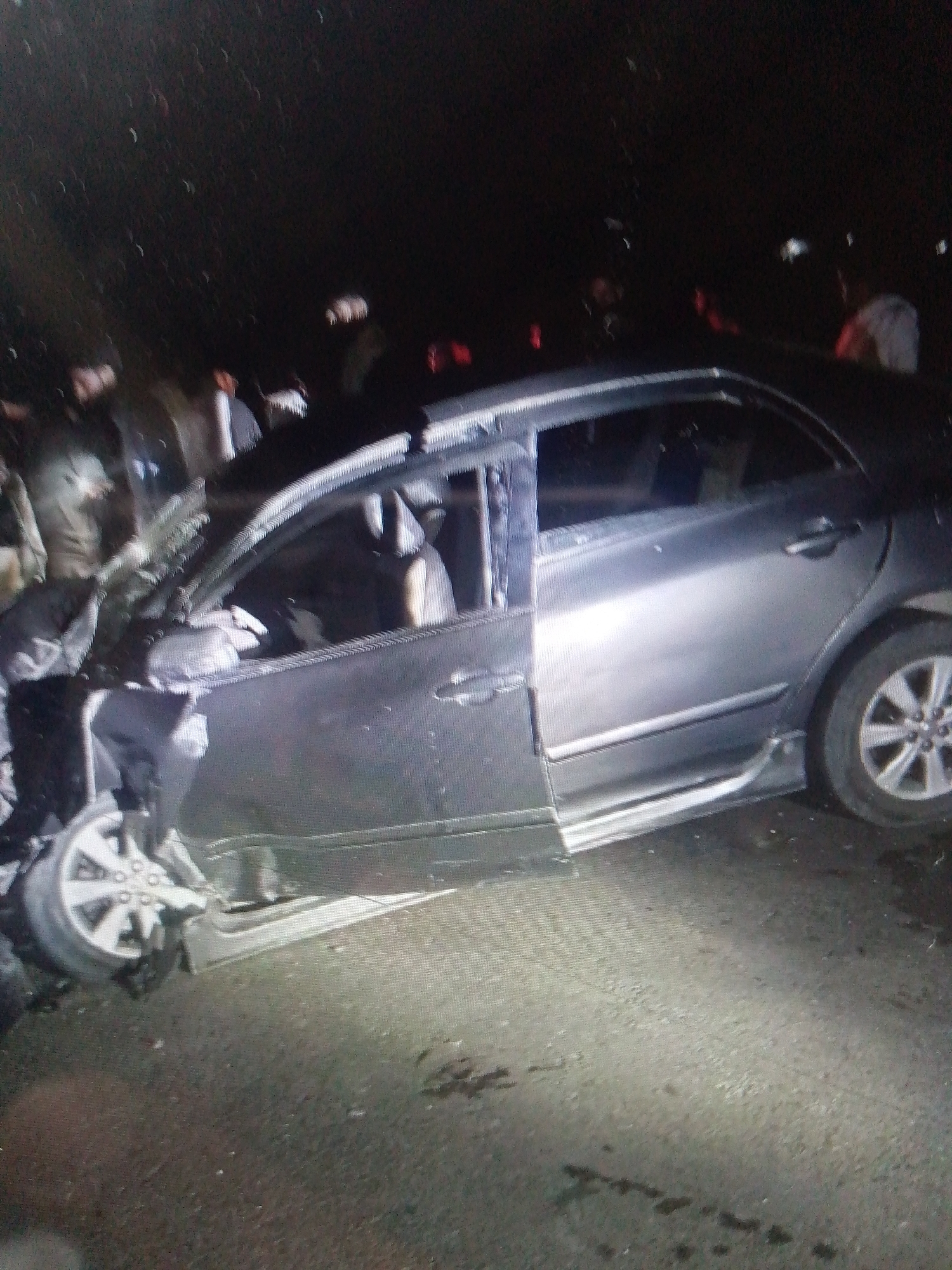 The image shows a car crash scene at night. The car, a gray sedan, has severe front-end damage, with the front wheel detached and significant deformation of the body. The roof and driver's side door are also damaged. People are gathered around the wrecked car. The location appears to be on a paved road, possibly in a city, but the specific details of the location (city, street name) are unknown. There are dark stains on the ground, potentially related to the accident.