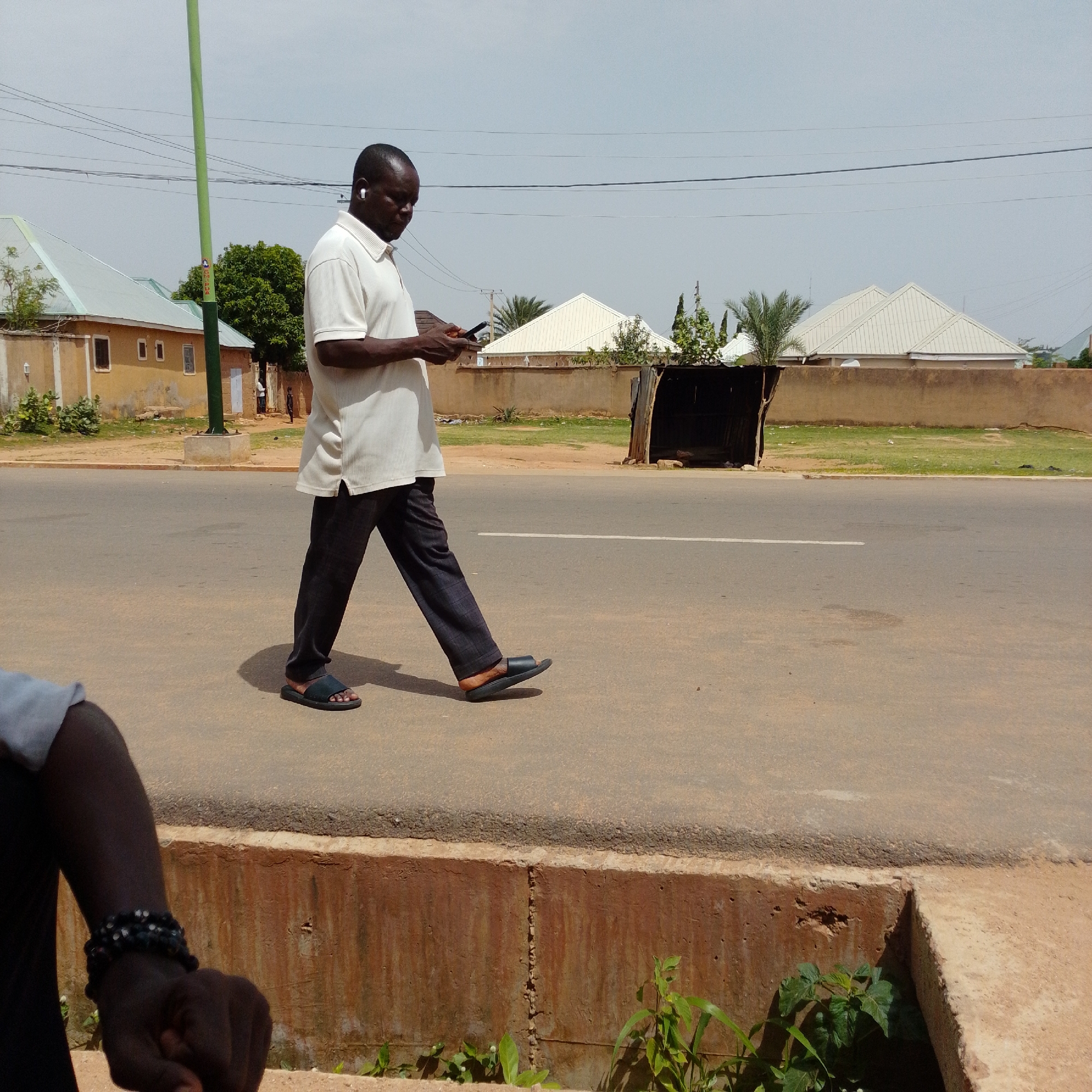In Gombe, Nigeria, a man walks on the side of a paved road. He is wearing a white polo shirt and dark trousers. He has earbuds in his ears and is looking at his phone. The road is bordered by a concrete sidewalk. In the background, there are several buildings with light-colored roofs, palm trees, and other vegetation. The sky is overcast.