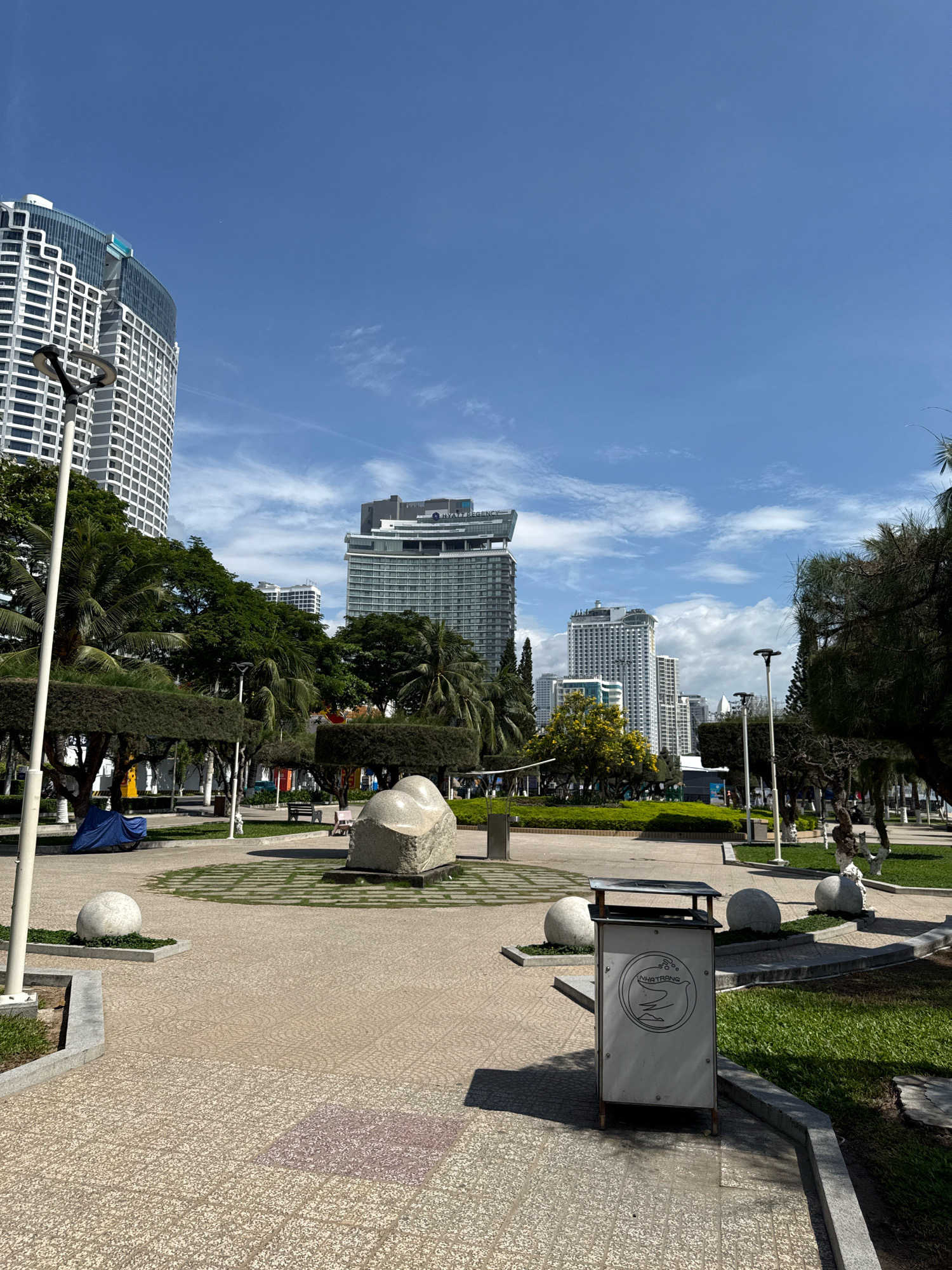 This is an outdoor, eye-level shot of a public park in Nha Trang, Vietnam, on a clear, sunny day with a few wispy clouds. The foreground features a textured, light-colored paved walkway. To the right of the walkway is a white trash can with a circular logo that appears to be a bird or swan design, and text below it that reads "NHATRANG". Further to the right, a patch of green grass is visible, bordered by a gray concrete curb.
In the midground, several large, smooth, light-colored spherical sculptures are placed on small patches of greenery, with a larger, irregularly shaped, light-colored stone sculpture in the center of a paved area. A bench and a pink object that might be another bench or a decoration are visible behind the large stone sculpture. Trees with dark green foliage are strategically placed throughout the park, some appearing to be trimmed into boxy shapes. A lamppost stands tall on the left side of the walkway. In the far left of the midground, a blue tarp covers a motorcycle or similar vehicle.
The background is dominated by a cityscape with several tall, modern buildings. The building closest to the left has a curved facade with a pattern of white horizontal and vertical lines. Further back, other high-rise buildings with glass windows and various architectural styles are visible. The sky is a vibrant blue. The overall scene depicts a well-maintained urban park designed for public recreation, situated in a coastal city.
