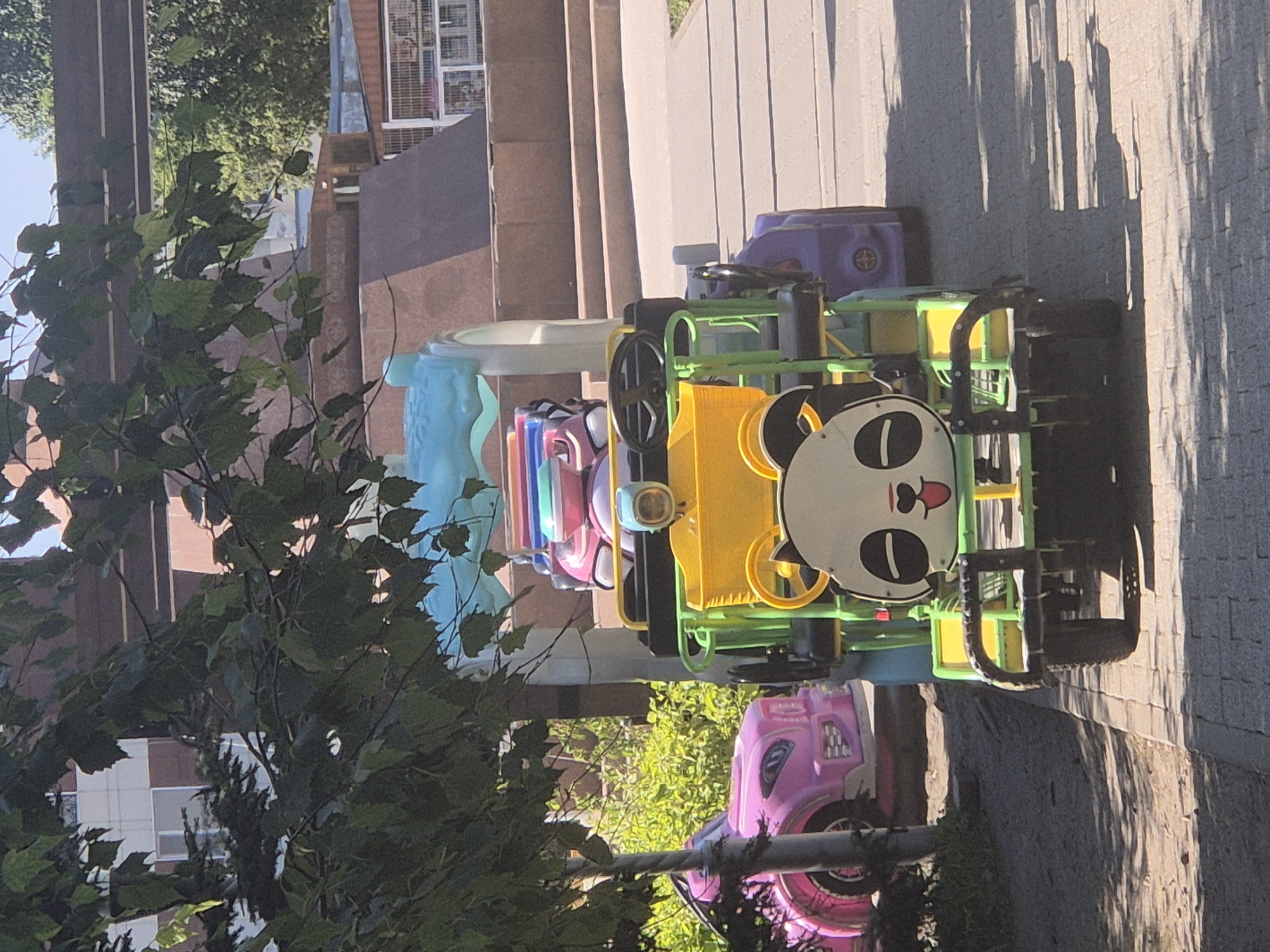In Kokshetau, Kazakhstan, on a sunny day, a vibrant children's amusement ride shaped like a panda-faced vehicle is parked on a paved walkway. The ride is primarily green and yellow, with a large panda face featuring black eyes and mouth, and a pink tongue. It has a blue canopy overhead. To the left of the ride, a pink toy car is visible, partially obscured by foliage. The background shows a multi-tiered stone structure and trees, suggesting a park or recreational area. The sun casts distinct shadows on the ground. No people are present in the immediate vicinity of the amusement ride.