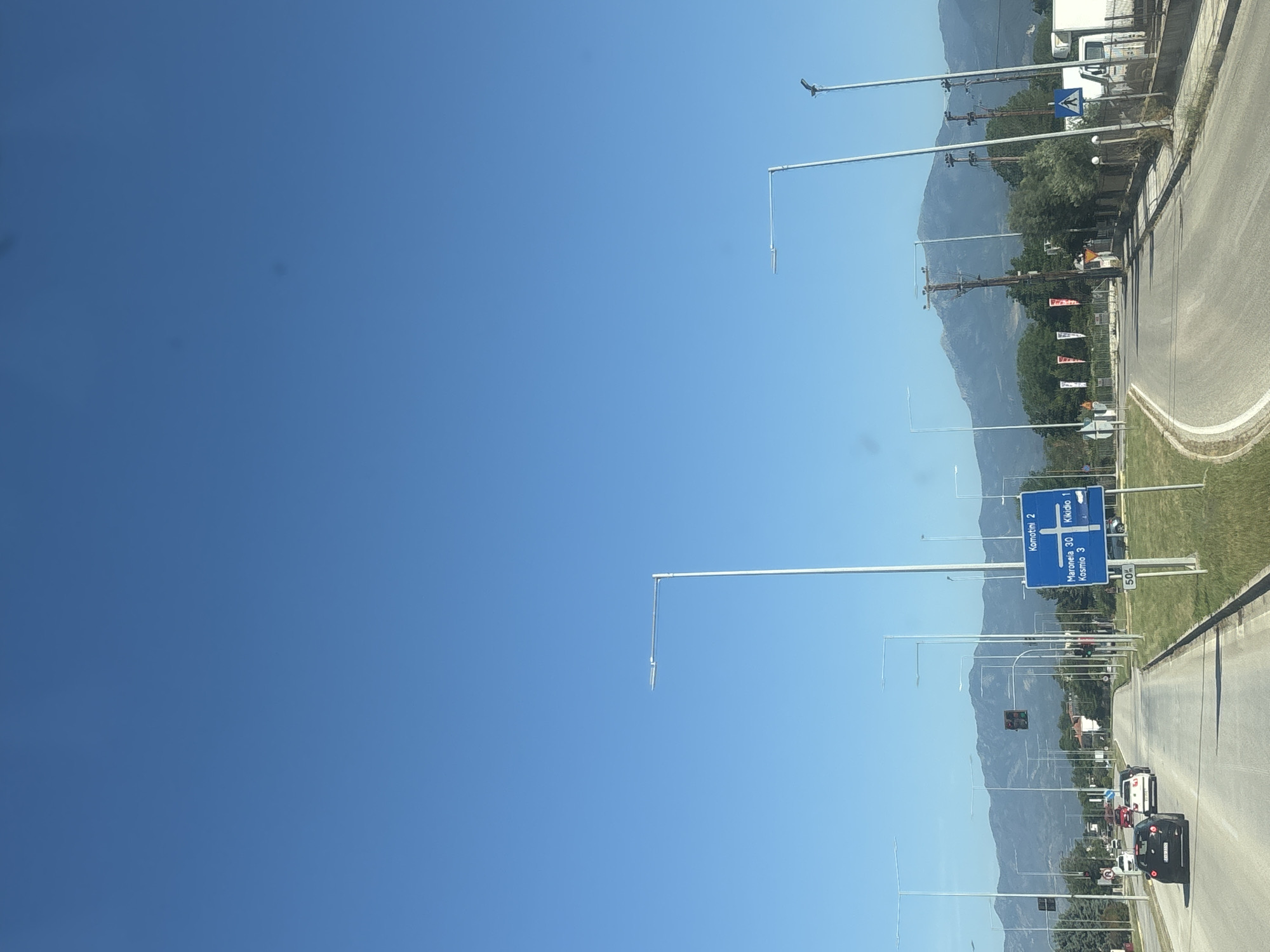 The image captures a wide, straight road in Kikidio, Greece, bordered by green grass and trees, with distant mountains under a clear blue sky. Several cars are driving on the road, and prominent blue traffic signs with white text indicate directions to Komotini, Maroneia, and Kikidio itself. Tall, modern streetlights line the road. In the background, a white truck and a smaller white vehicle are parked. The scene appears to be during daylight, likely morning or afternoon.