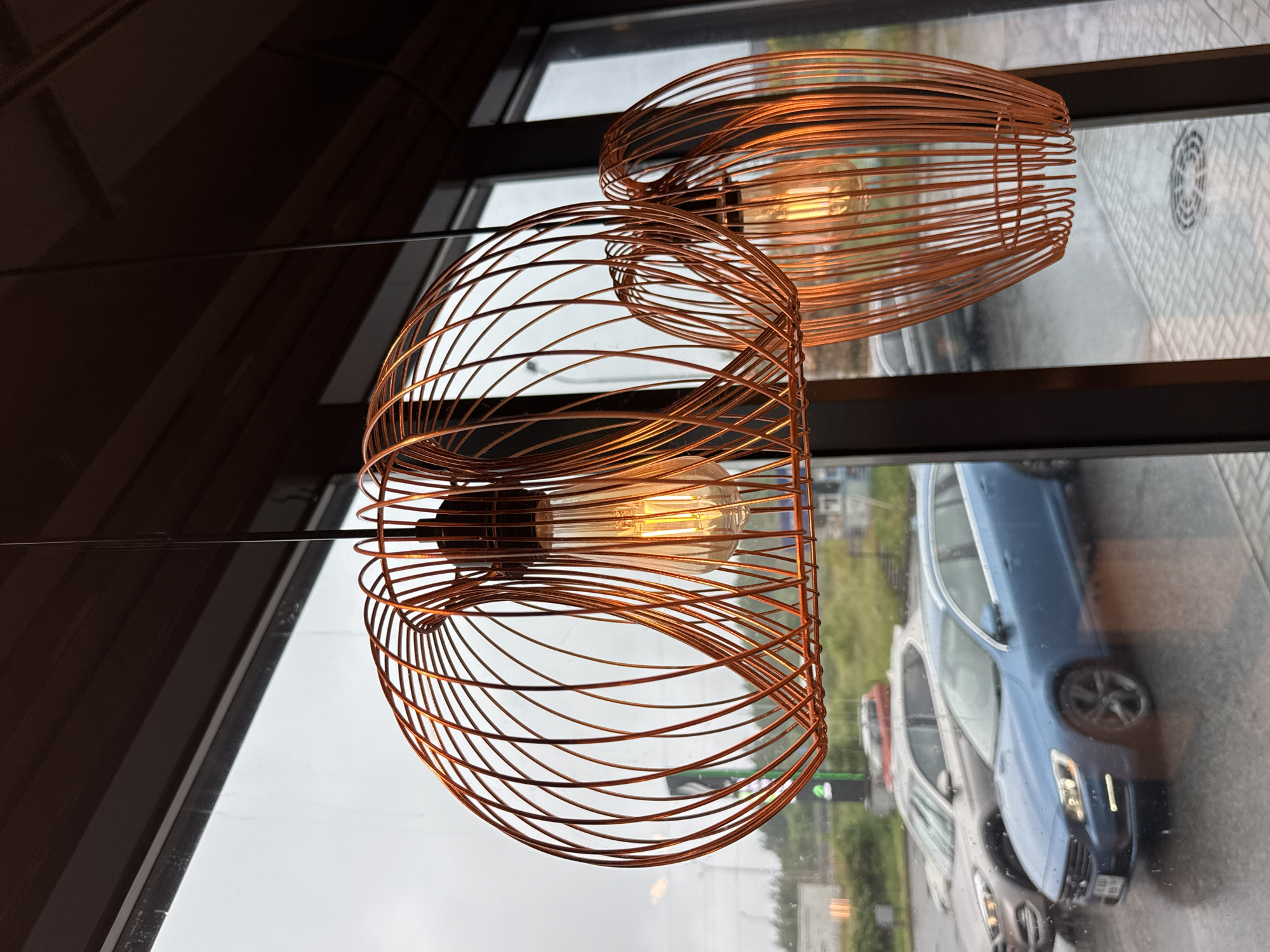 Two copper wire pendant lights hang in front of a large window. The lights have an abstract, cage-like design and emit a warm glow from visible Edison bulbs. Through the window, a rainy day scene is visible. Several cars are parked on a wet asphalt surface, with a blurred background of greenery and buildings. One prominent car is a blue sedan. The overall mood is somewhat moody and atmospheric due to the overcast weather and indoor lighting. The image appears to be taken from inside a building, likely a cafe or restaurant, looking outwards.