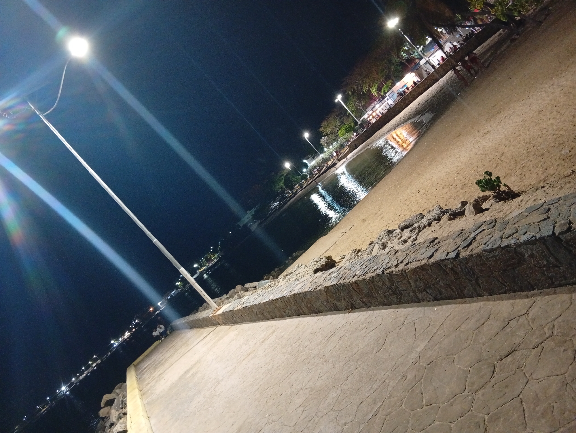 The image captures a night scene in Juan Griego, Venezuela. In the foreground, there's a textured, light-colored pavement. A low wall constructed of dark stones separates the pavement from a sandy beach. Beyond the beach, there's water reflecting the lights of buildings and street lamps. Several streetlights illuminate the beach and pavement. In the distance, the lights suggest the presence of buildings and establishments along the shoreline. Some people are visible in the distance near what appears to be a building with open storefronts. A small plant is visible on the beach near the low stone wall.