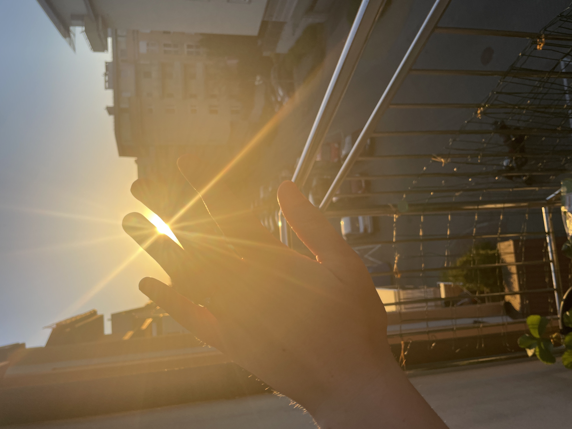 The image shows a hand held up towards a bright sun, with visible sunbeams radiating from the space between the fingers. The background includes buildings and a railing, suggesting a balcony or similar elevated location. The buildings appear to be in an urban setting. The lighting is intensely bright due to the direct sunlight.