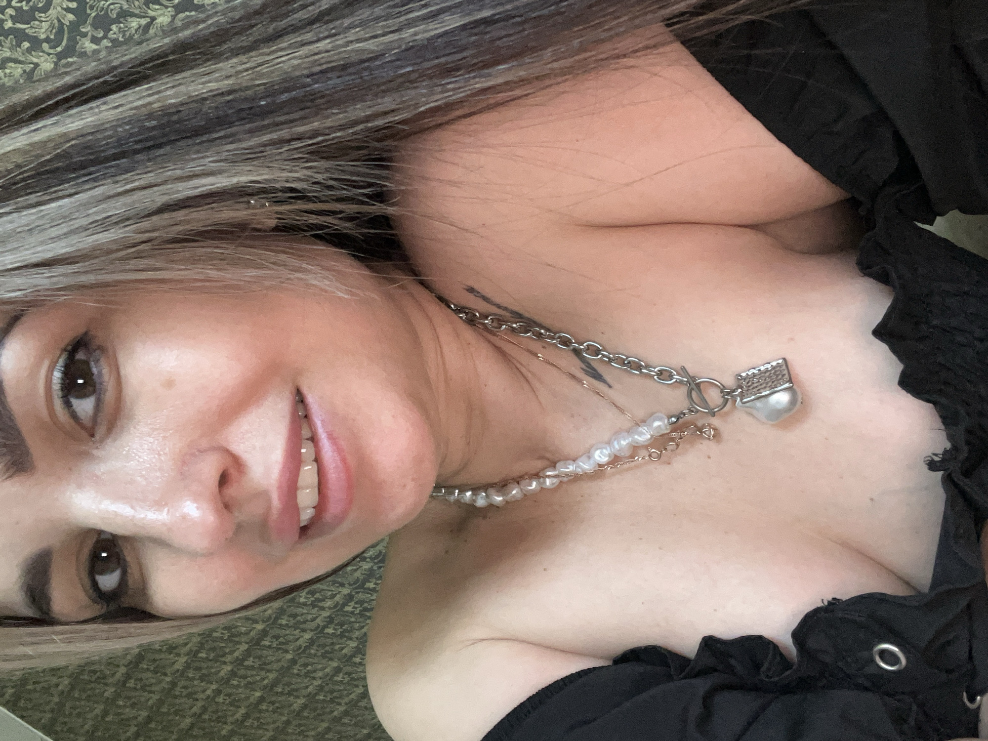 A medium shot depicts a woman with light skin and a complexion suggesting middle age, her face tilted to the left. Her hair is dyed with dark roots fading into lighter, blonde streaks. Her eyebrows are dark and well-defined, while her eyes are dark brown. Her lips are full, with visible teeth. She is wearing a black, off-the-shoulder top that is textured. A layered necklace with pearls and a silver charm adorns her neck. A tattoo is visible above her collarbone. The background is a textured green surface. The location is Unknown, Unknown.