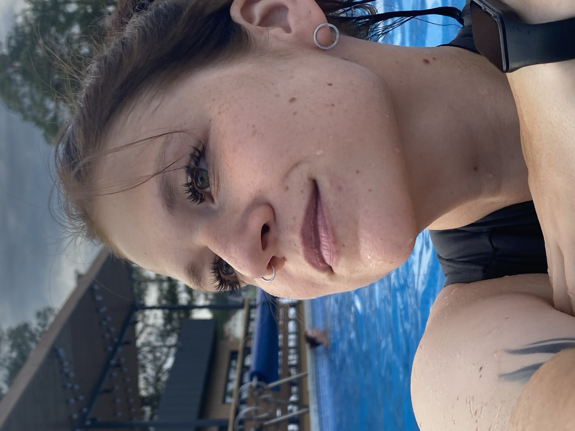 A close-up, profile view captures a young woman with freckles, green eyes, and dark hair styled in a messy bun, likely at an outdoor pool facility in Votkinsk, Russia. She is partially submerged in the bright blue water, her skin glistening with droplets. Her gaze is directed upwards and to the left, with a subtle smile on her lips. She wears a nose ring and a circular earring in her left ear, and a black smartwatch on her right wrist. Her left shoulder and arm, bearing a dark tattoo, are visible. In the background, the blue water of the pool extends, with another person visible in the distance. A covered structure with lights is present above the pool. The sky above is cloudy and overcast.