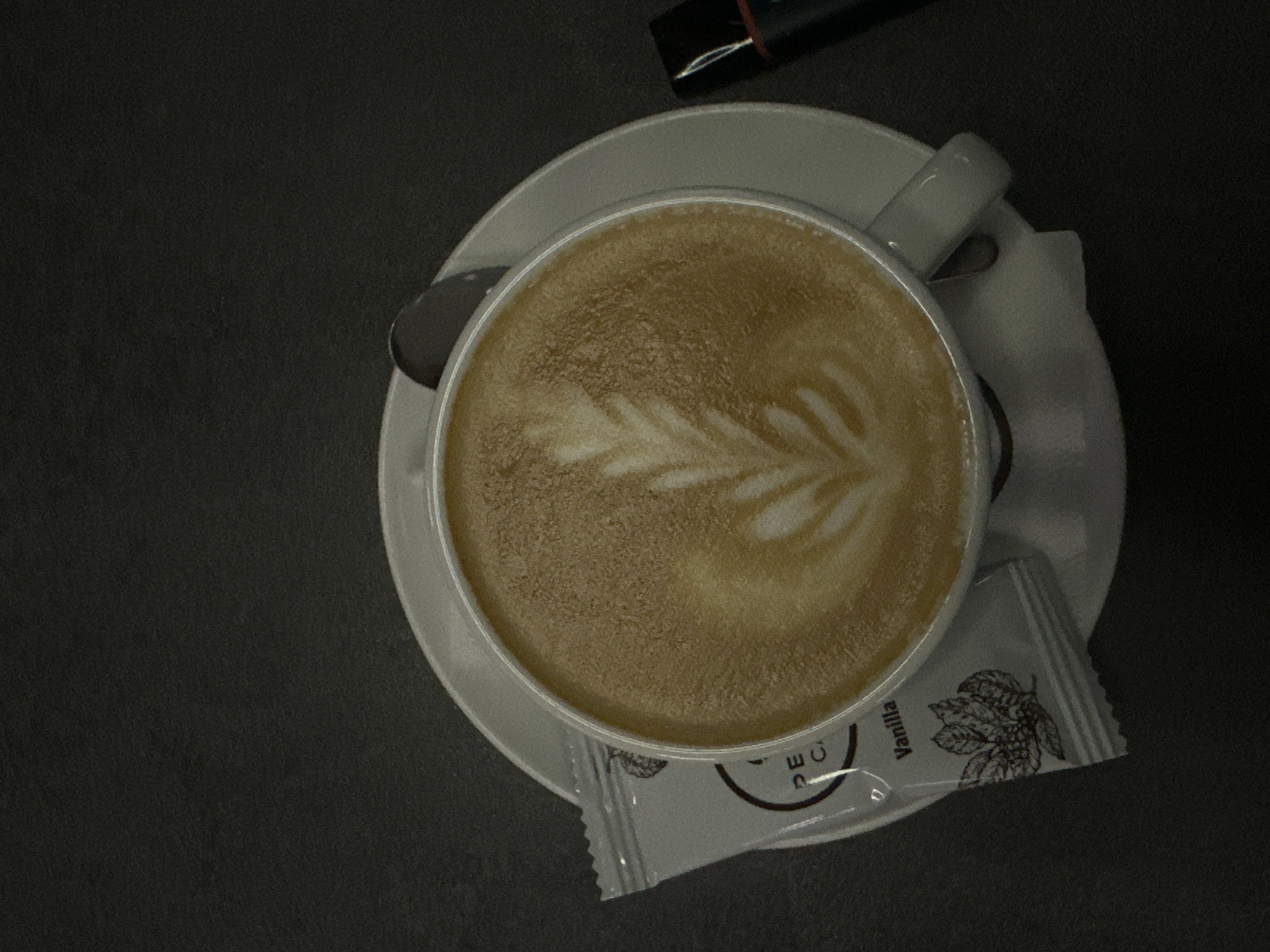 The image shows a cappuccino with latte art, served on a white saucer with a spoon and a vanilla sugar packet, likely in a cafe in Stara Zagora, Bulgaria. A black object resembling a pen or marker is also present near the cup. The background is a dark, neutral surface, suggesting a table. The scene is dimly lit, creating a subtle contrast.