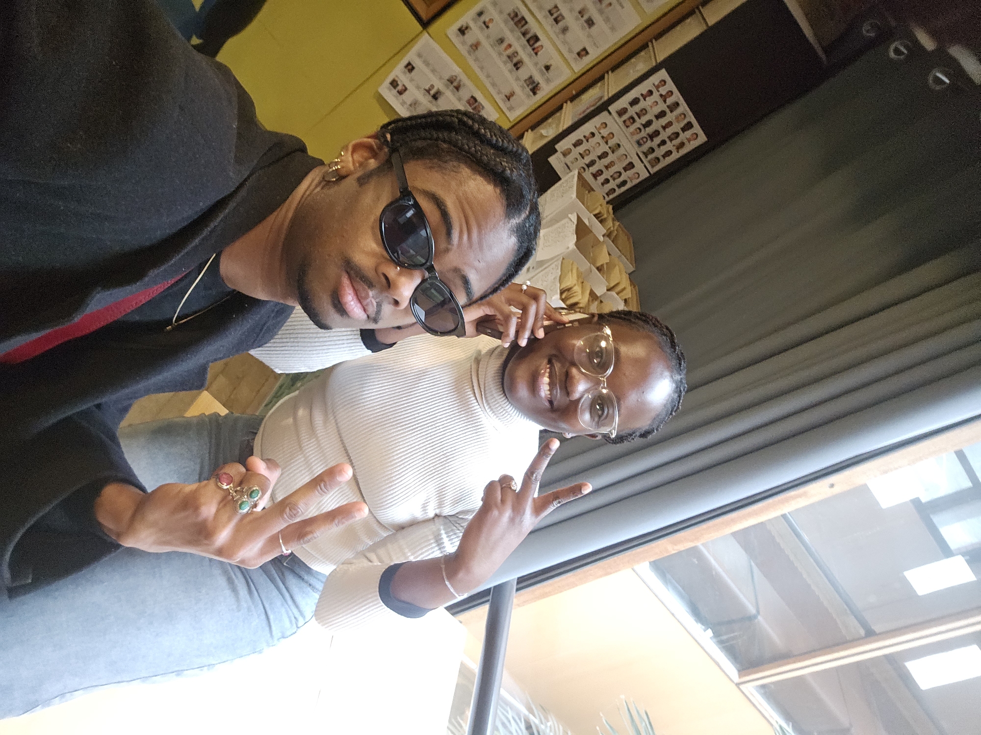 Two individuals are captured in a selfie-style photograph. The person in the foreground, presumed to be male, is wearing sunglasses and a dark jacket over a red garment. Gold earrings are visible. The second person, presumed to be female, is positioned slightly behind and to the side. She is wearing glasses and a light-colored turtleneck sweater. Both individuals are displaying the peace sign with their hands. The setting appears to be indoors, possibly a cafe or restaurant, judging by the interior design and the presence of what looks like a menu board in the background. The backdrop includes a window with a view to the outside. Though not explicitly visible in the image, there is no information that it is set in Paris, France.