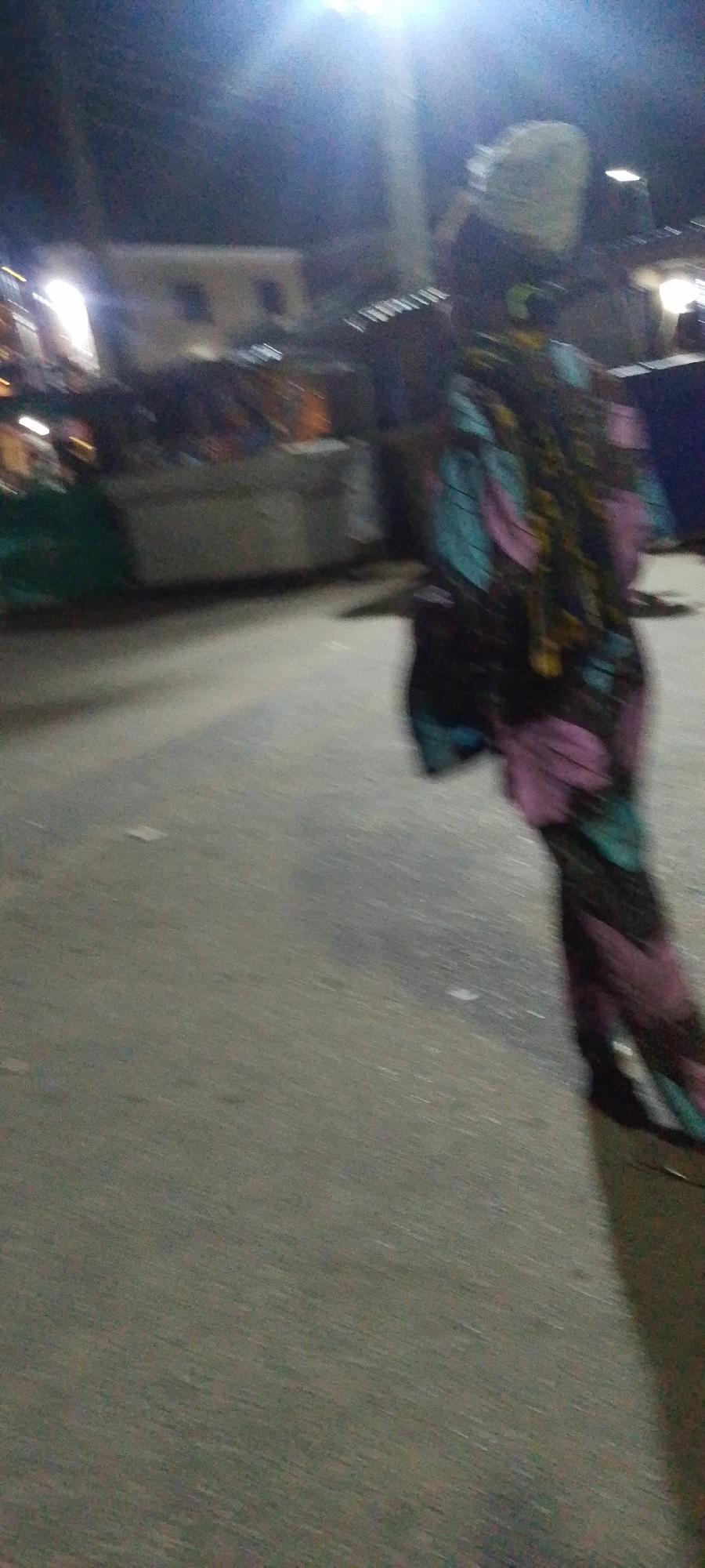 A blurry nighttime scene in Lokoja, Nigeria, featuring an individual in patterned clothing walking on a street. Buildings and lights are visible in the background, creating a sense of urban activity.