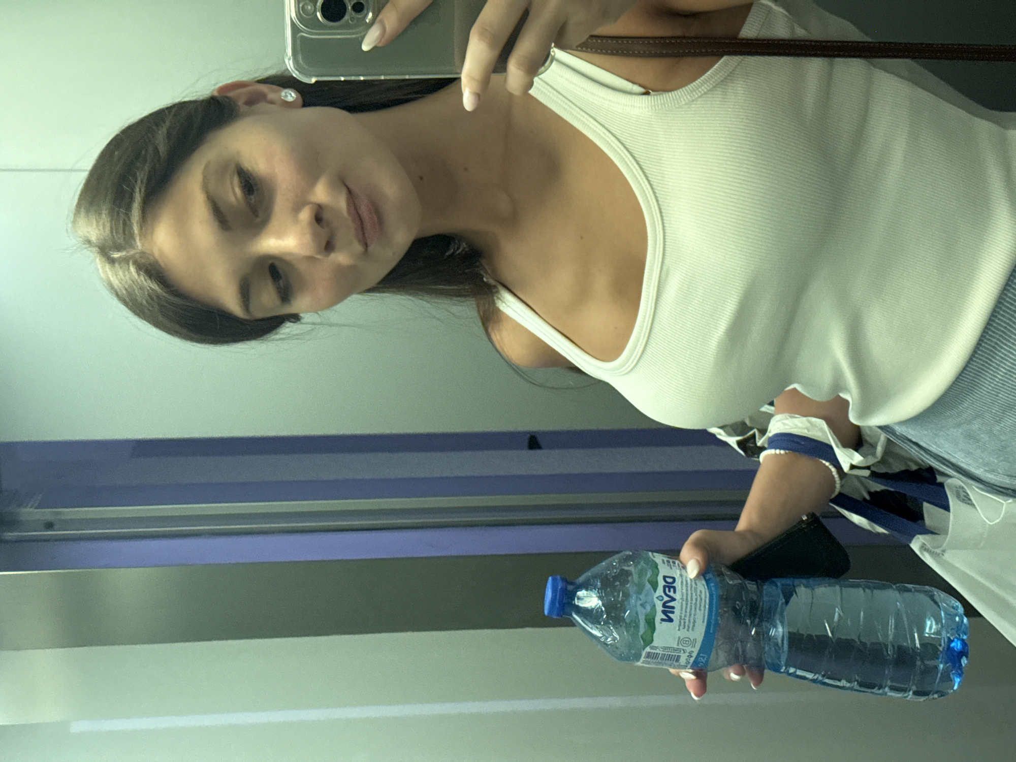 A young woman with long brown hair and light skin is taking a selfie in an elevator. She is wearing a white tank top and blue jeans, and holding a bottle of water in her left hand. She is also wearing a silver earring in her right ear and a pearl bracelet on her left wrist. The elevator appears to be in a modern building, with grey walls and a blue vertical stripe on the left side. There are no visible distinguishing features to indicate the specific location within Burgas, Bulgaria.