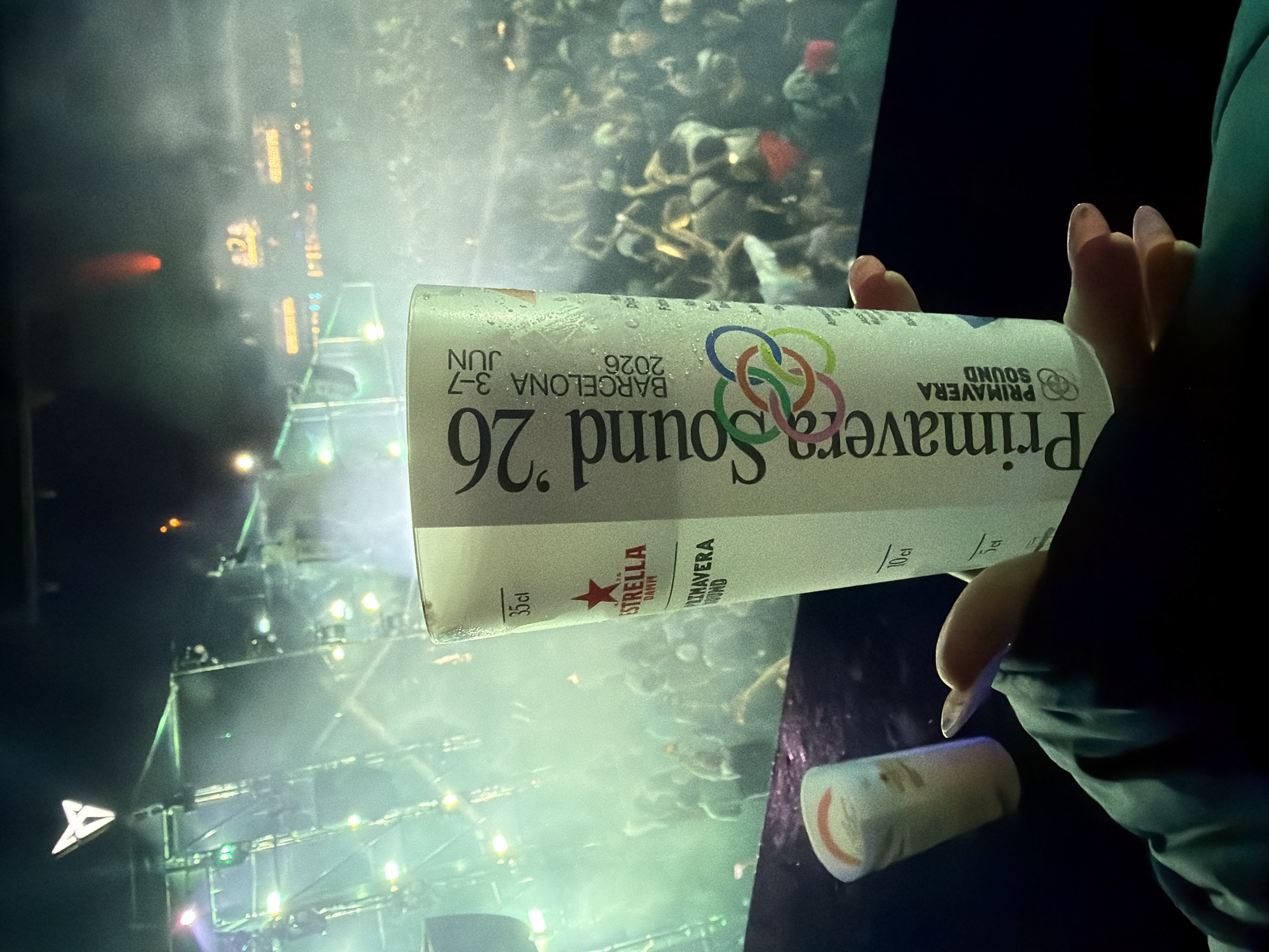 This low-angle, first-person photograph captures a night scene at the Primavera Sound festival, held at Parc del Fòrum in Sant Adrià de Besòs, Spain. In the immediate foreground, a left hand with light-colored fingernail polish, partially obscured by a dark teal sleeve, holds a white reusable festival cup. The cup displays condensation and is printed with "Primavera Sound 26", "Barcelona 3-7 JUN 2026", the "Estrella Damm" logo featuring a red star, and measurement markings for 35cl, 10cl, and 5cl, along with a colorful interlocking rings logo. To the left of the main cup, a smaller, white cylindrical object with a red/orange rim rests on a dark, possibly wet, surface. The midground features a dense, blurred crowd of people, illuminated by green stage lights and atmospheric fog. In the background, a large metal stage scaffolding structure is visible, emitting bright white and green lights. A prominent, illuminated white arrow-shaped logo (resembling the Cupra brand emblem) is affixed to the upper left of the stage structure. Further in the distance, another blurred, illuminated sign with yellow text, possibly reading "24," is discernible through the fog.