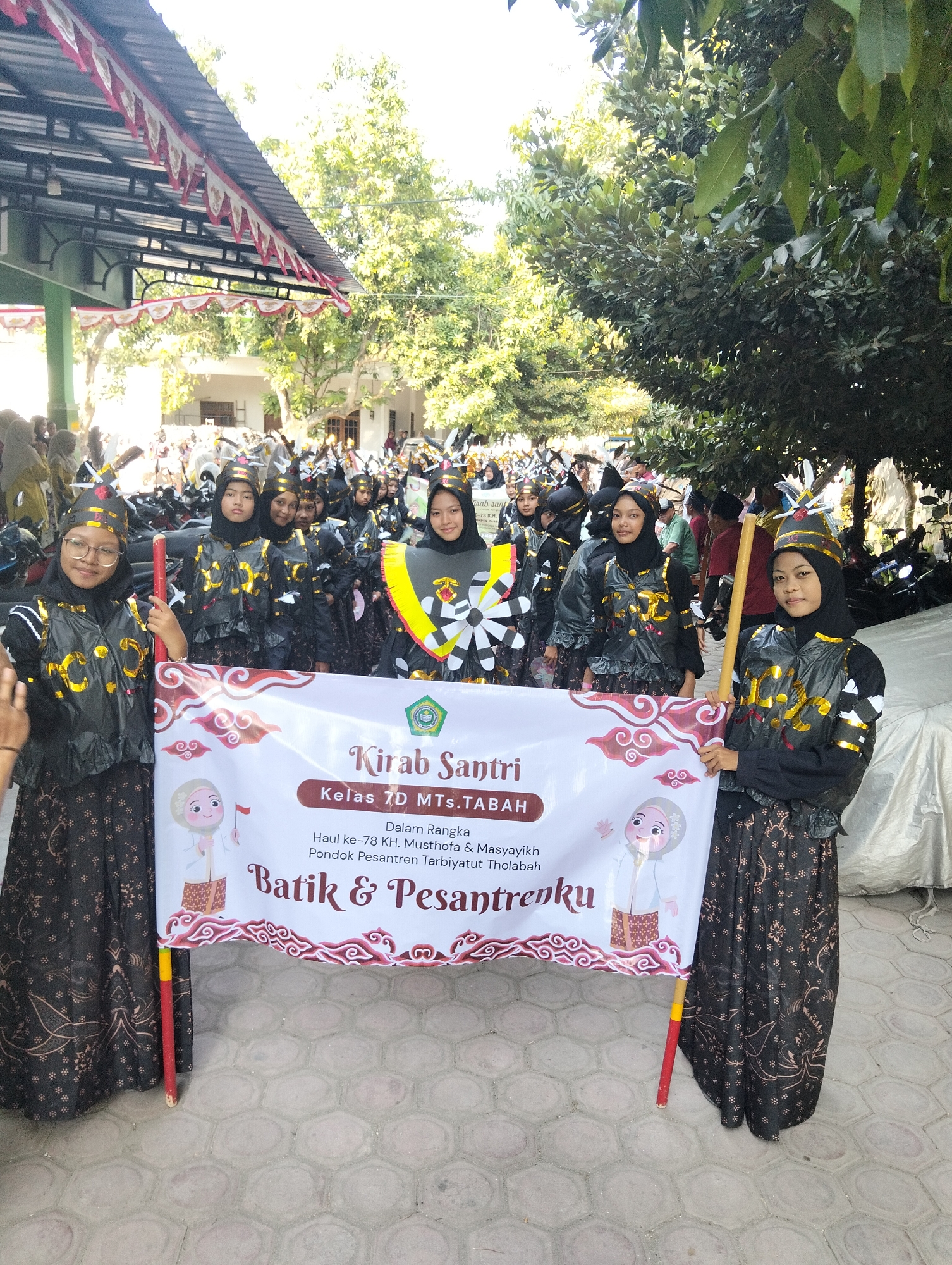 In Kranji, Indonesia, a group of young women, identified as students of Kelas 7D MTs. TABAH, are participating in a parade. They are dressed in matching black and gold uniforms, including headscarves, vests, and patterned skirts. Some wear decorative hats. The students are holding a banner that reads "Kirab Santri" and "Batik & Pesantrenku," indicating a cultural event celebrating Islamic boarding schools and batik. The banner also commemorates the 78th anniversary of Musthofa & Masyayikh Pondok Pesantren Tarbiyatut Tholabah. The parade appears to be taking place outdoors, with trees and buildings visible in the background, suggesting an urban or semi-urban setting. The ground is paved with hexagonal tiles. The overall atmosphere seems festive and organized.