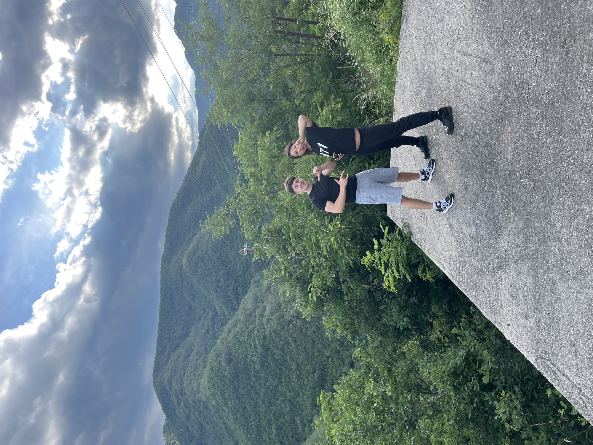 Two young adults, a male and a female, are posing for a photo on a concrete platform. The male, on the left, is wearing a black t-shirt and grey shorts, with white and black sneakers. He is making a hand gesture with his right hand and holding his left hand up. The female, on the right, is wearing a black t-shirt with "371" printed on it and black sweatpants, with black sneakers. She is holding her right hand up, forming a peace sign, and her left hand is also raised. They are standing on a sloped concrete surface with a backdrop of lush green mountains and trees under a dramatic sky with clouds and sunlight breaking through. The location is the city of Pechinska, Bulgaria.