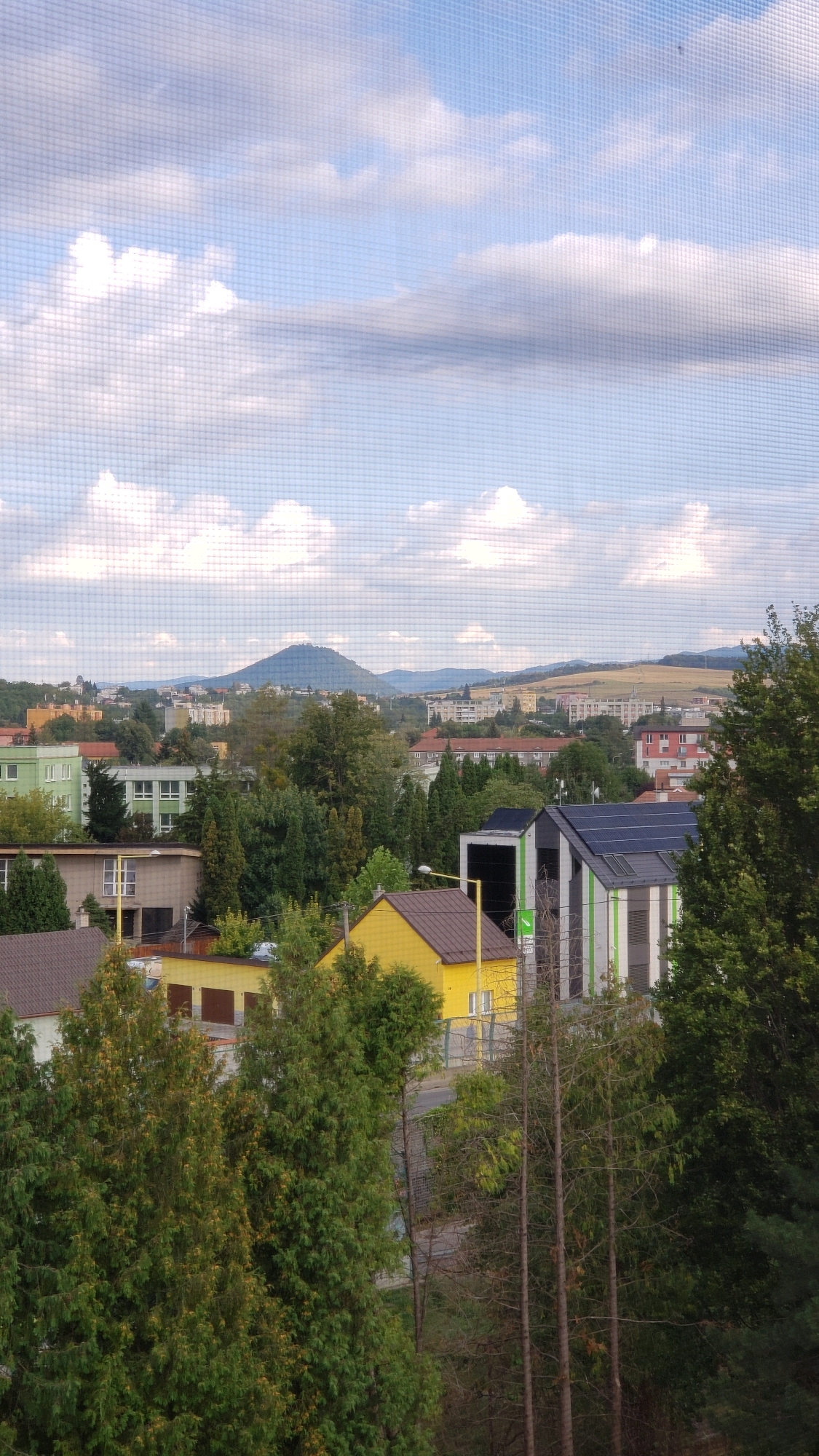 The image depicts a suburban landscape with a view of a distant conical mountain, possibly Solivarsky vrch, from the city of Prešov, Slovakia. The foreground is dominated by dense green trees, including evergreens and deciduous trees. In the middle ground, several buildings are visible. One is a bright yellow house with a brown roof. Adjacent to it is a modern-looking building with a multi-colored facade, featuring vertical green and white stripes, and a black section that may be a solar panel array. Another building with a brown roof and white window frames is also present. Further back, more residential and industrial buildings can be seen, along with a prominent green building. The background reveals a range of hills and mountains under a partly cloudy sky. The image appears to be taken through a screen, indicated by the grid pattern superimposed on the scene. There are no people or animals visible in the image. The overall impression is one of a quiet residential area on a clear day with a picturesque natural backdrop.