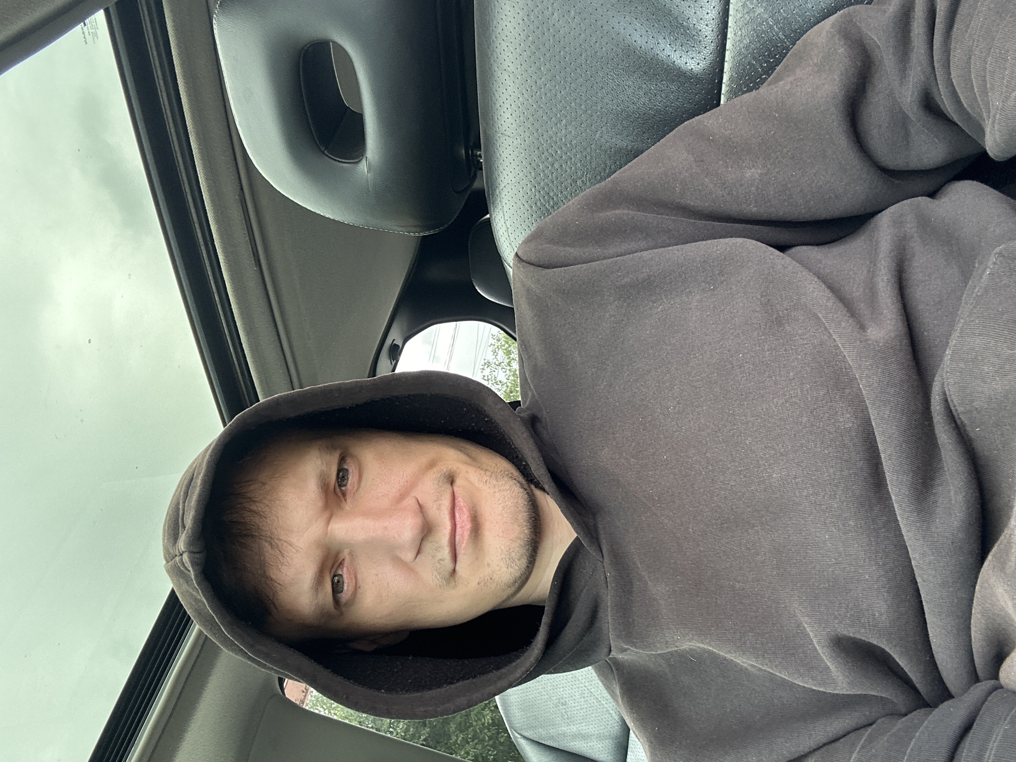 A man in a dark hoodie sits in the driver's seat of a car, looking forward. The car has a sunroof, which is open and allows light to filter in from an overcast sky. The man's face is framed by the hood of his sweatshirt. The interior of the car is visible, including the black leather headrest of the passenger seat and part of the dashboard. The image appears to have been taken from a smartphone, as indicated by the selfie perspective. The overall lighting suggests it is daytime, though the sky is cloudy. There is no indication of the specific location in the city of Makiv, Ukraine.
