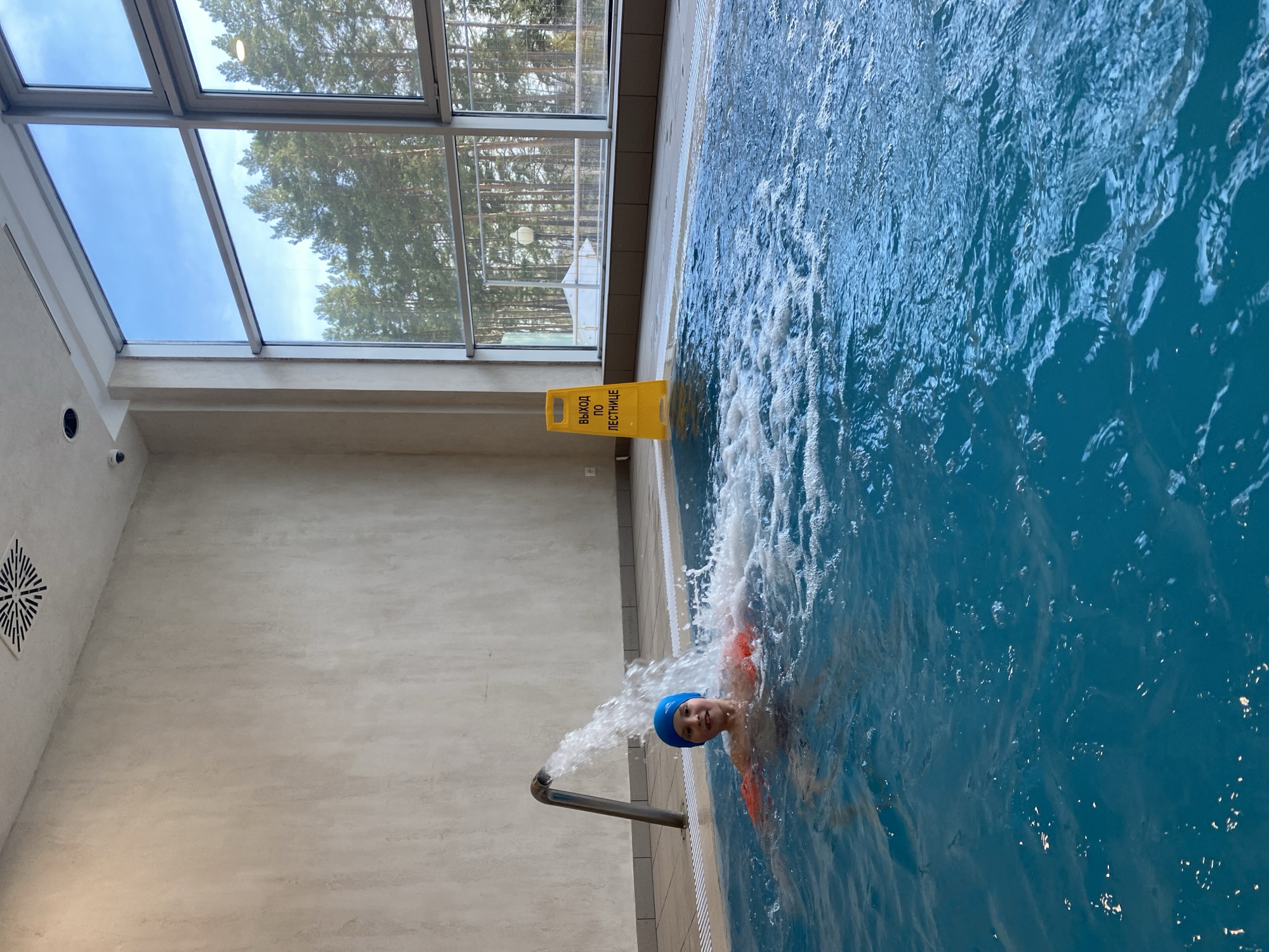 A medium, high-angle shot depicts a person with a blue swim cap in a swimming pool. The pool's water is a vibrant blue. The person has orange floaties on their arms and appears to be near a water feature that shoots water into the pool.
The pool is indoors, with a large window providing a view of trees, possibly a forest or park, outside. A yellow warning sign stands near the edge of the pool, displaying text in Russian, likely related to a staircase. The walls and decor have a modern, clean aesthetic.
The location is possibly the "Tyumen" hot springs water park in the city of Tyumen, Russia, based on architectural style.