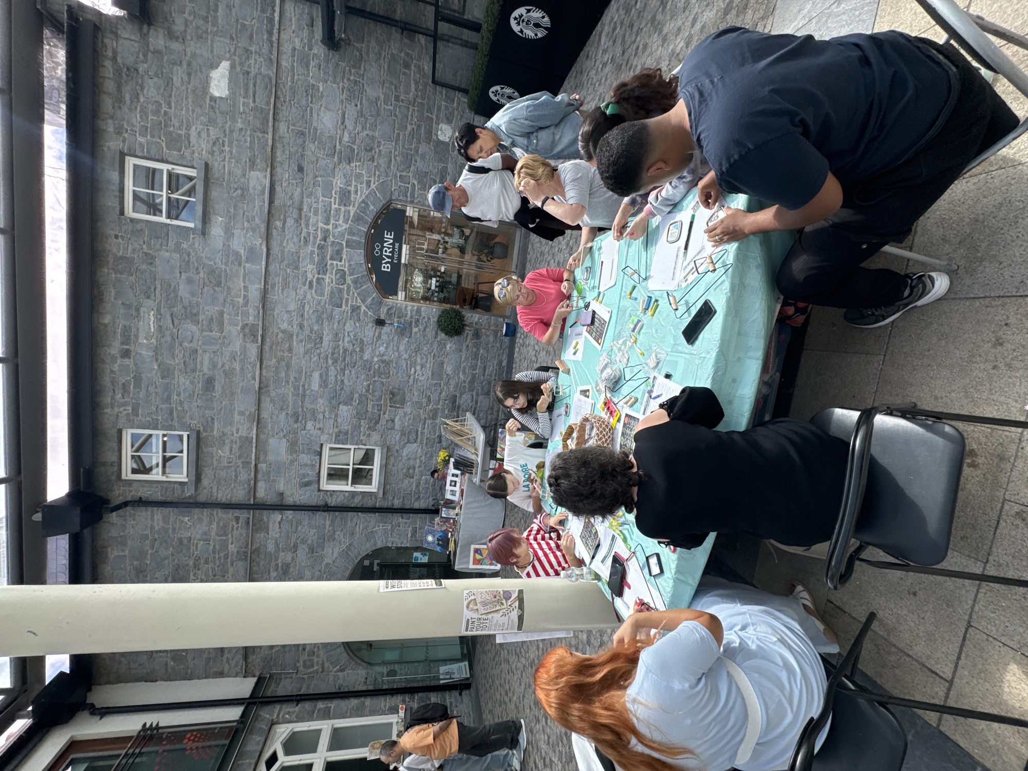 The image depicts an outdoor craft activity taking place in Kilkenny, Ireland. A group of people are gathered around a large, round table covered with a light blue tablecloth. The table is laden with art supplies, including papers, pens, and craft materials, suggesting a workshop or event. Several individuals, including adults and children, are actively engaged in crafting, with some focused on their creations and others interacting with each other. The backdrop features a distinctive stone building with arched doorways and windows, characteristic of Kilkenny's historic architecture. A sign above an entrance reads "BYRNE EYECARE," indicating a commercial establishment nearby. The scene appears to be set in a public square or courtyard, with paved flooring and surrounding buildings contributing to the urban setting. The overall atmosphere is one of community engagement and creative activity.