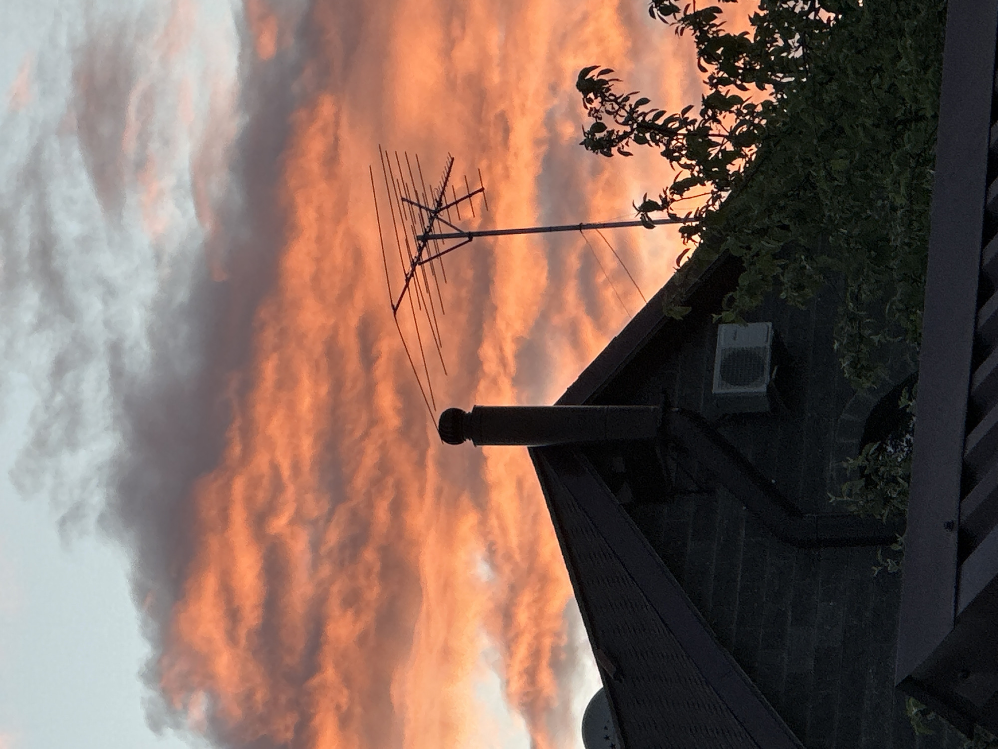 The image captures a sunset scene with clouds illuminated in shades of pink and orange. In the foreground, there's a building with a dark roof. A chimney and a TV antenna are attached to the roof. A patch of green foliage, possibly from a tree or bush, is partially visible against the sky. An air conditioner is visible on the side of the building. The photograph is taken from a low angle, looking upwards. There is no information to determine the exact location in Nizhny Novgorod, Russia. No people are visible in the image.