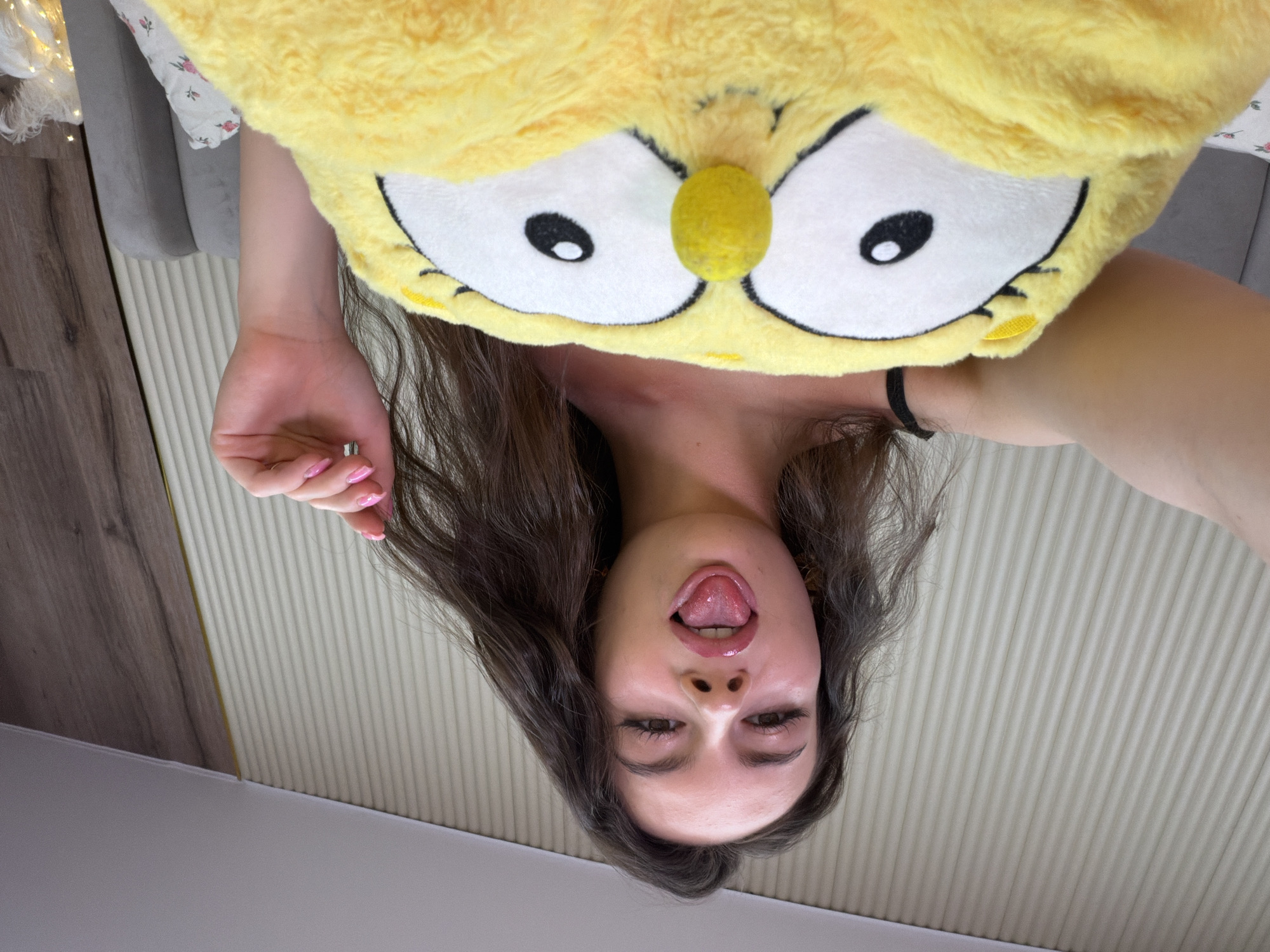 A young woman with long, dark hair is holding a large, yellow, owl-shaped plush toy in front of her. She is looking directly at the camera with her tongue sticking out. The background appears to be a bedroom, with a slatted wall and a headboard of a bed visible. The woman is wearing a black strap, possibly from a top. The image was likely taken indoors. There is no information about the city of Chaikovsky, Russia, in this image.
