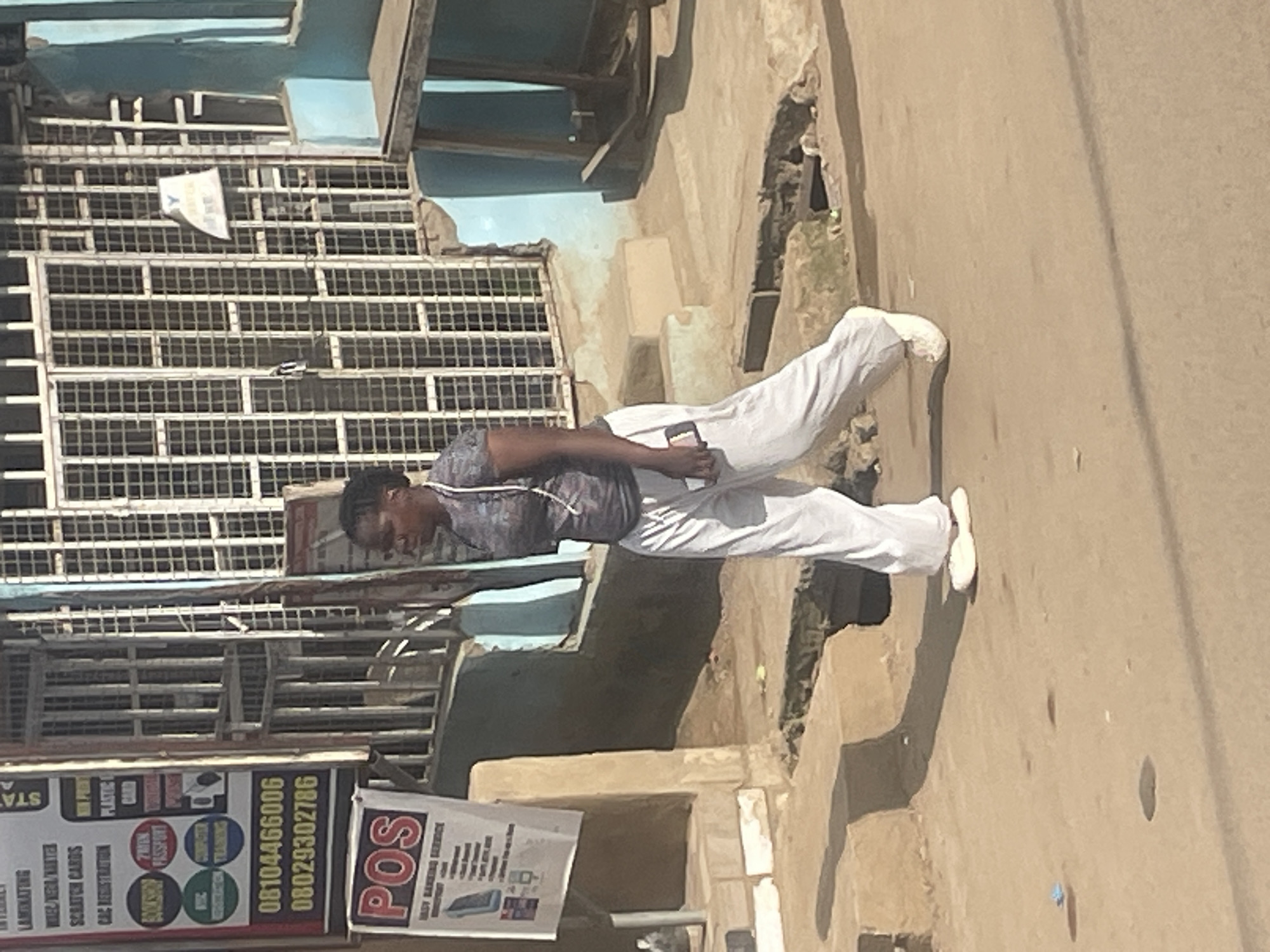 A young Black woman is walking from left to right across a sandy street in what appears to be an outdoor market or commercial area in Abuja, Nigeria. She is wearing a grey patterned t-shirt, loose white pants, and white slip-on shoes. She is holding a dark-colored cell phone in her right hand. The woman is walking past a storefront with metal bars on the windows and a sign advertising various services, including laminating, scratch cards, and CAC registration. A POS (Point of Sale) sign is visible on a white banner hanging from the storefront. The ground is unpaved and uneven, with some potholes and debris. In the background, there is a building with blue painted walls and a wooden structure that resembles a stall or table. The lighting suggests it is daytime.